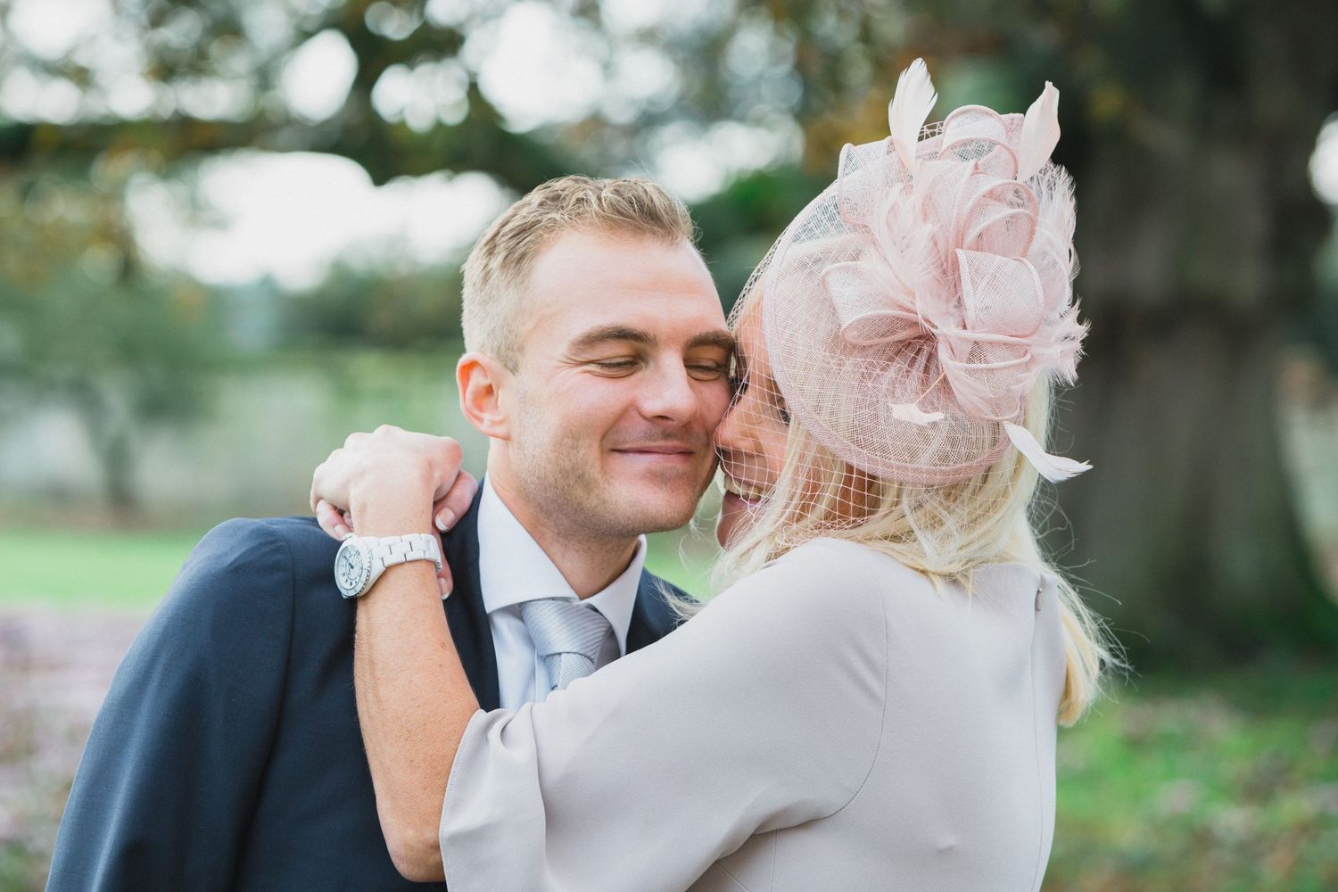 A couple embraces tenderly at a wedding, the bride wearing a pink fascinator and the groom in a suit with a light blue tie.