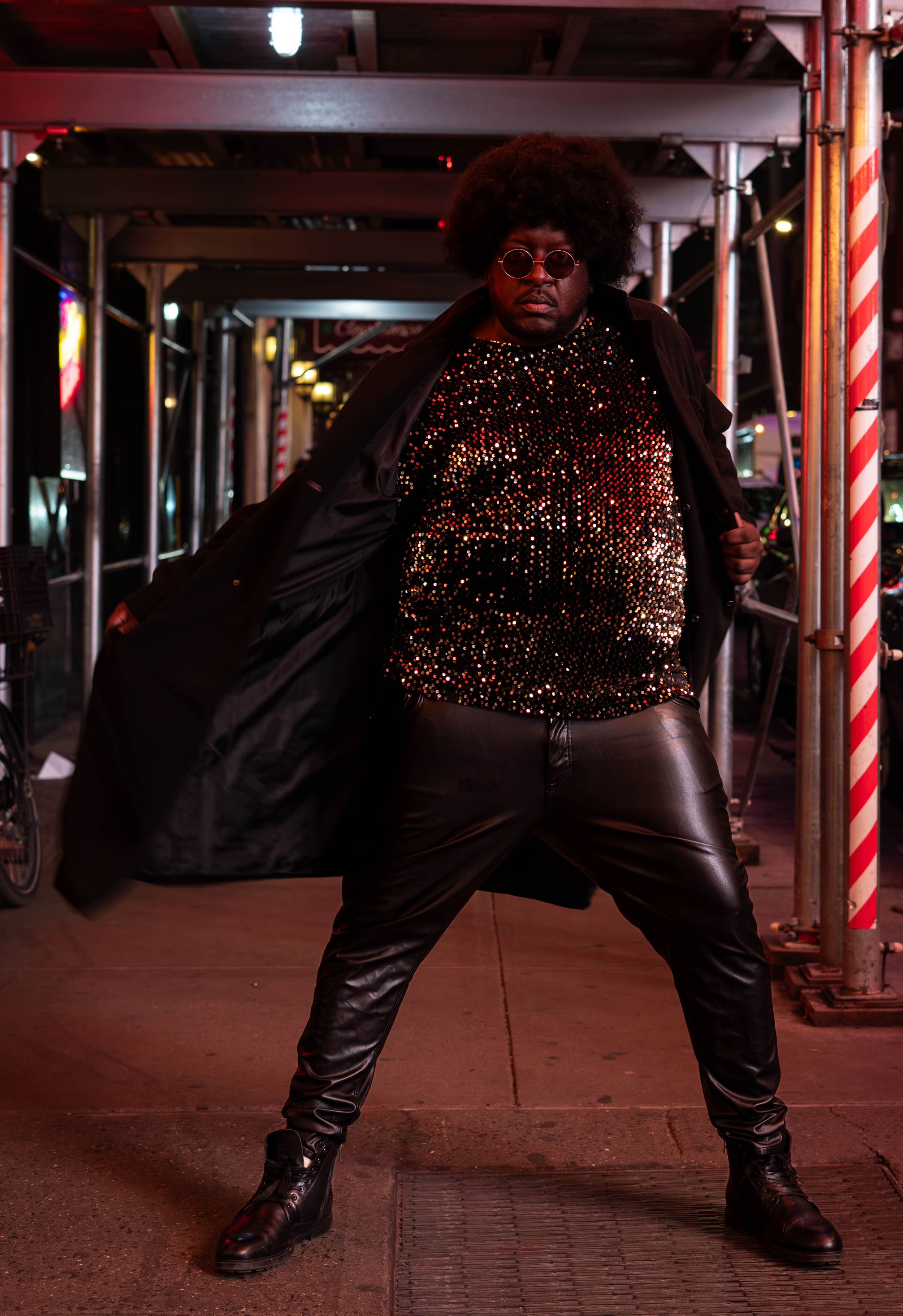Person in sequined top, black coat, leather pants, and afro wig poses confidently on urban street at night.