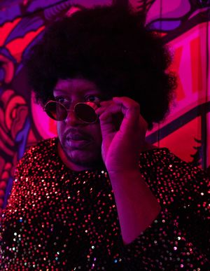 Person with afro hairstyle wearing round sunglasses and sequined top posed against colorful mural backdrop.
