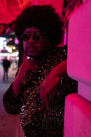 A stylish man with an afro and round sunglasses poses in a glittering sequin jacket under vibrant pink neon lights.