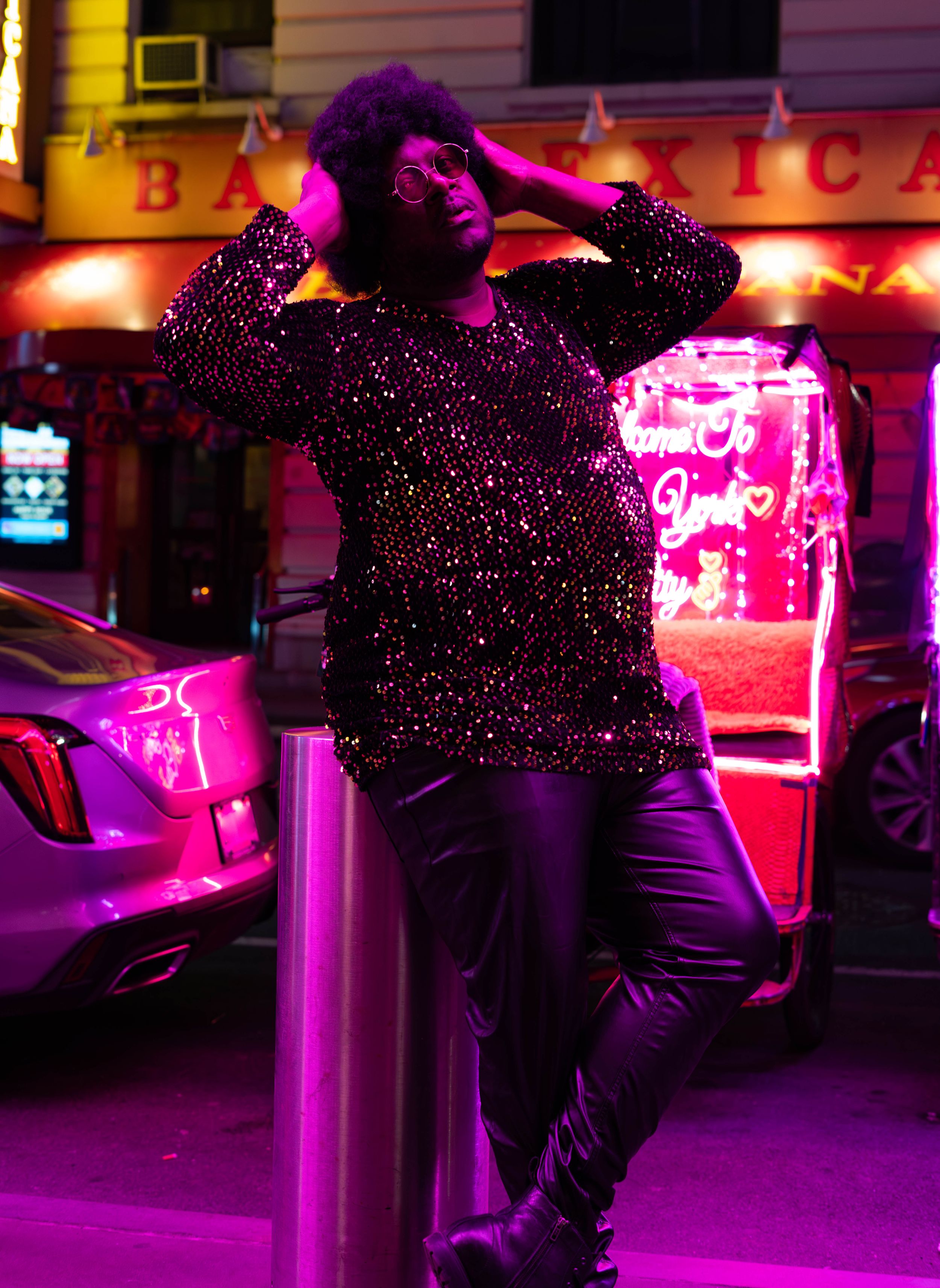 Person in sequin top and faux fur hat poses against bollard under vibrant pink neon lights outside a bar.