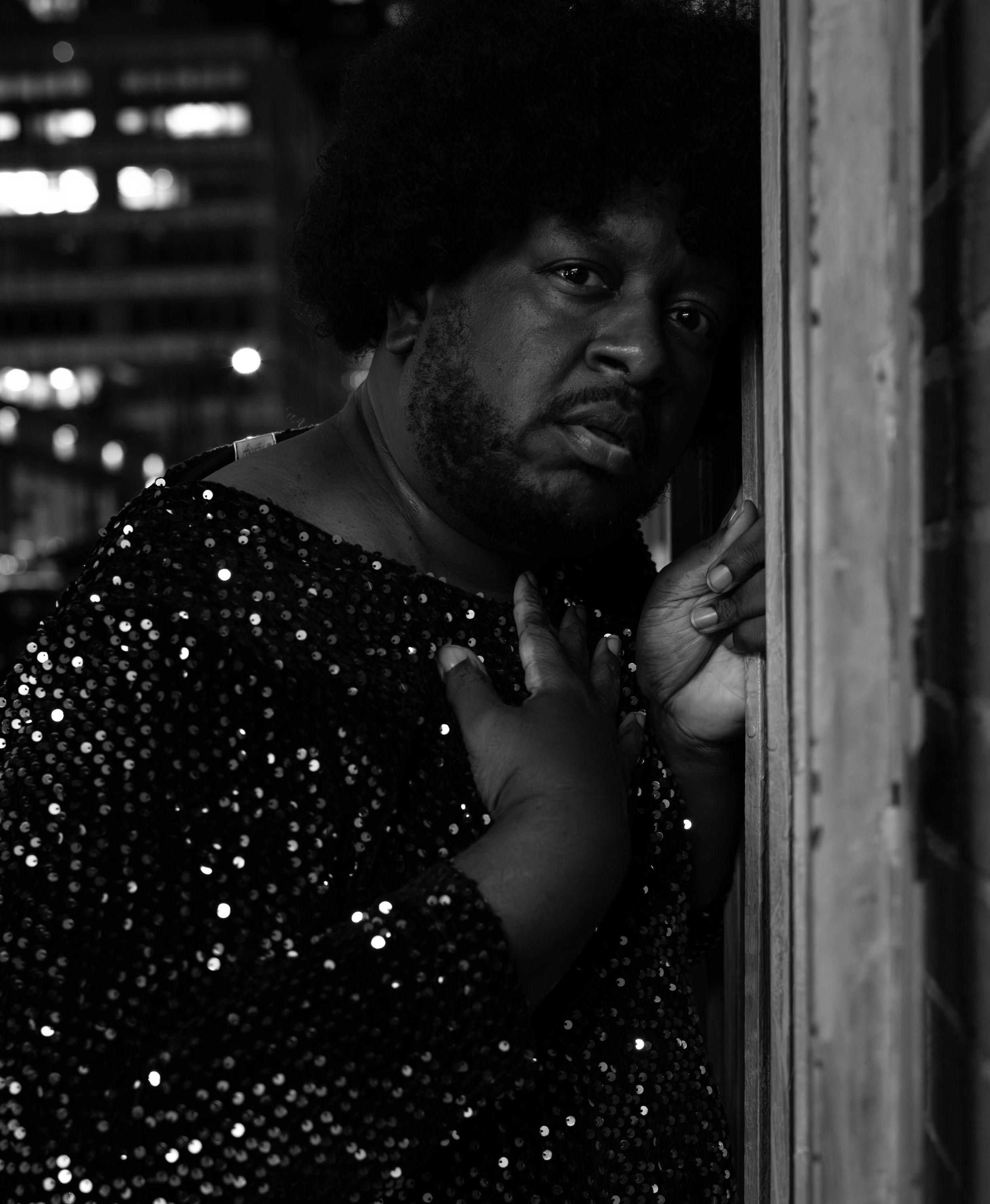 Black and white portrait of a person in sequined top leaning against a wooden frame at night.