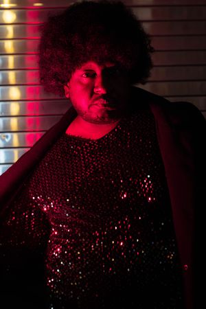 Person with afro hair wearing sequined top under red neon light in dark studio setting.