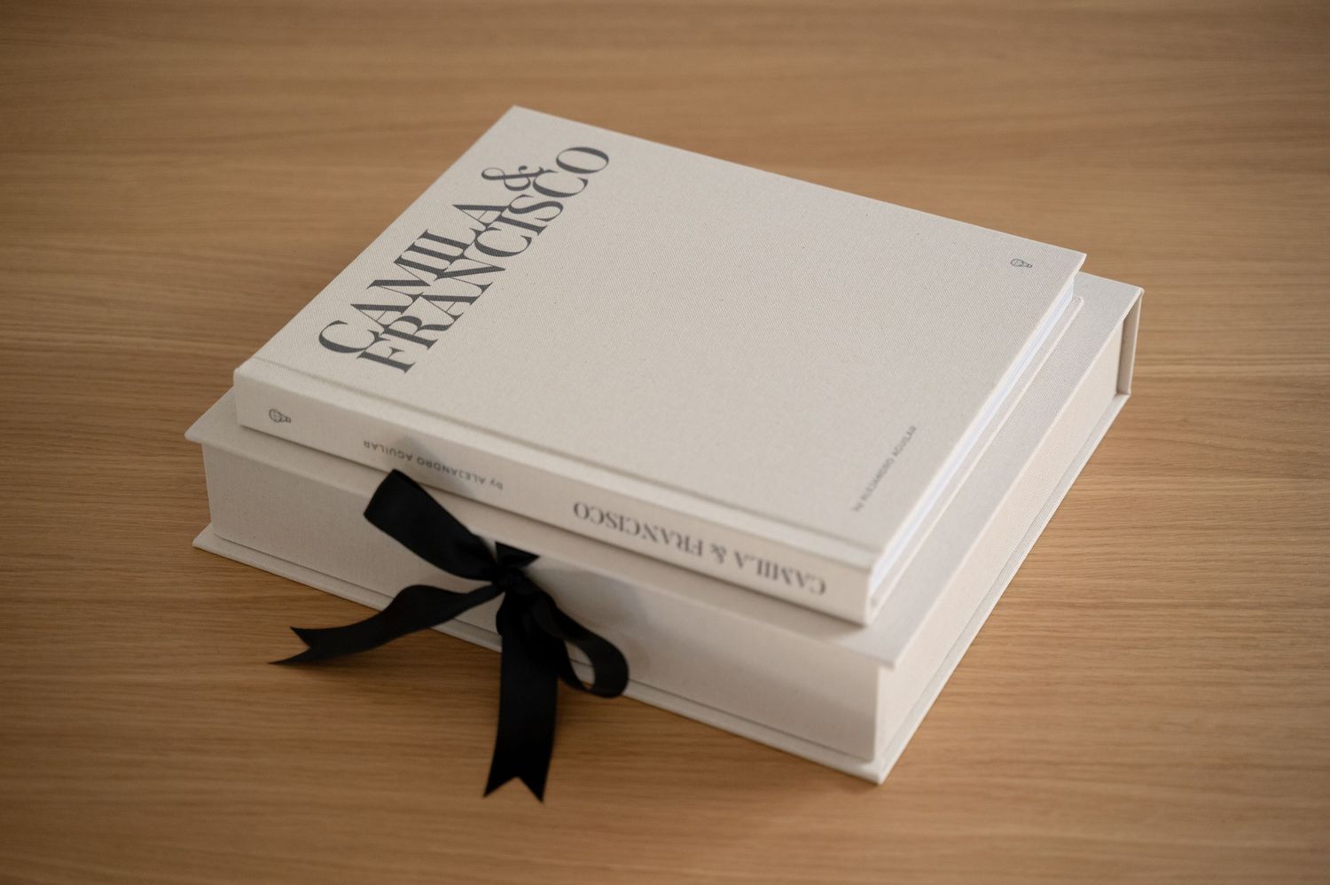 Elegant white wedding photo book with black ribbon bow, personalized with Camila & Francisco names on wooden surface.