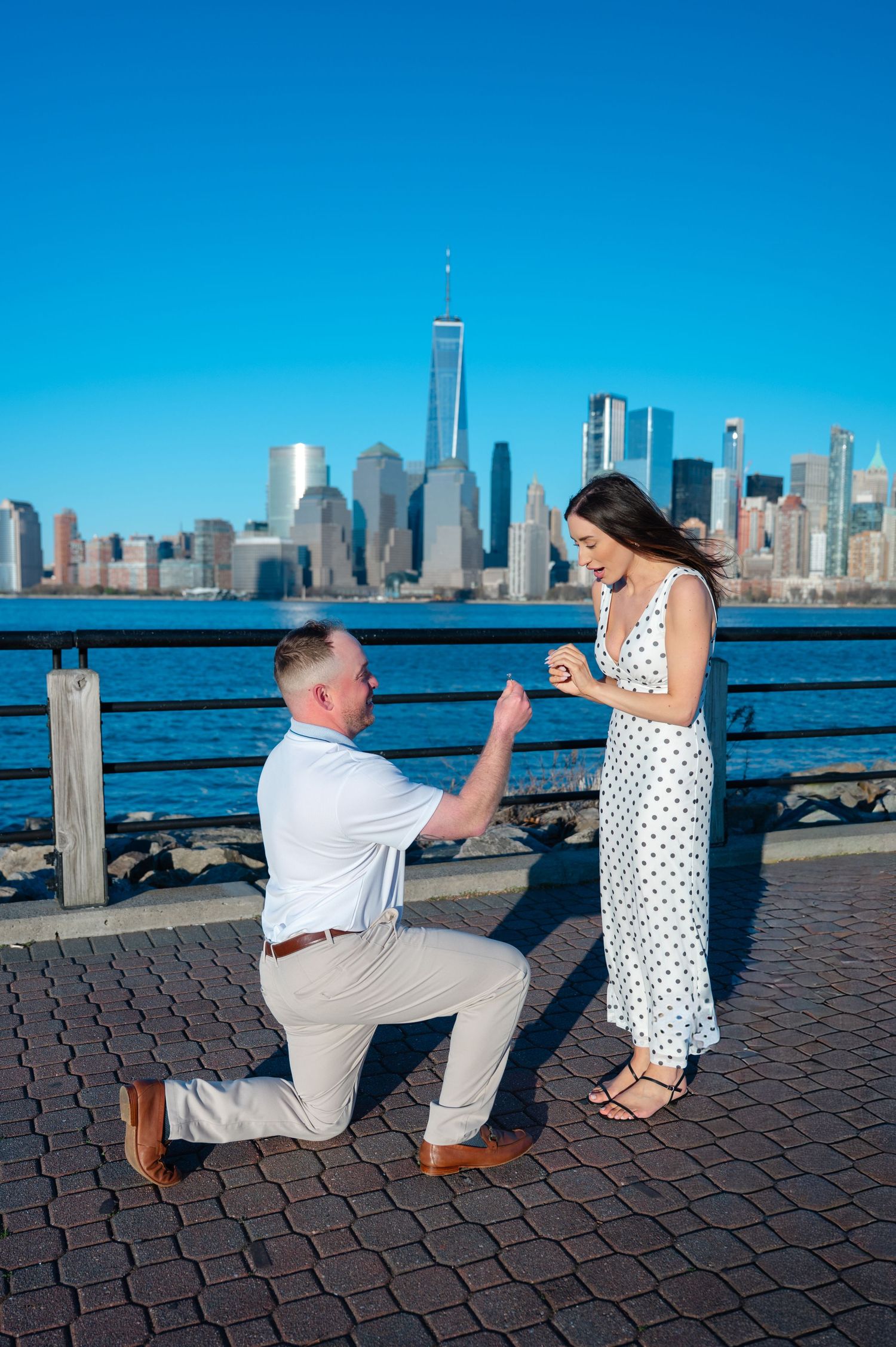 Man proposes on one knee to woman in polka dot dress with NYC skyline and One World Trade Center backdrop.
