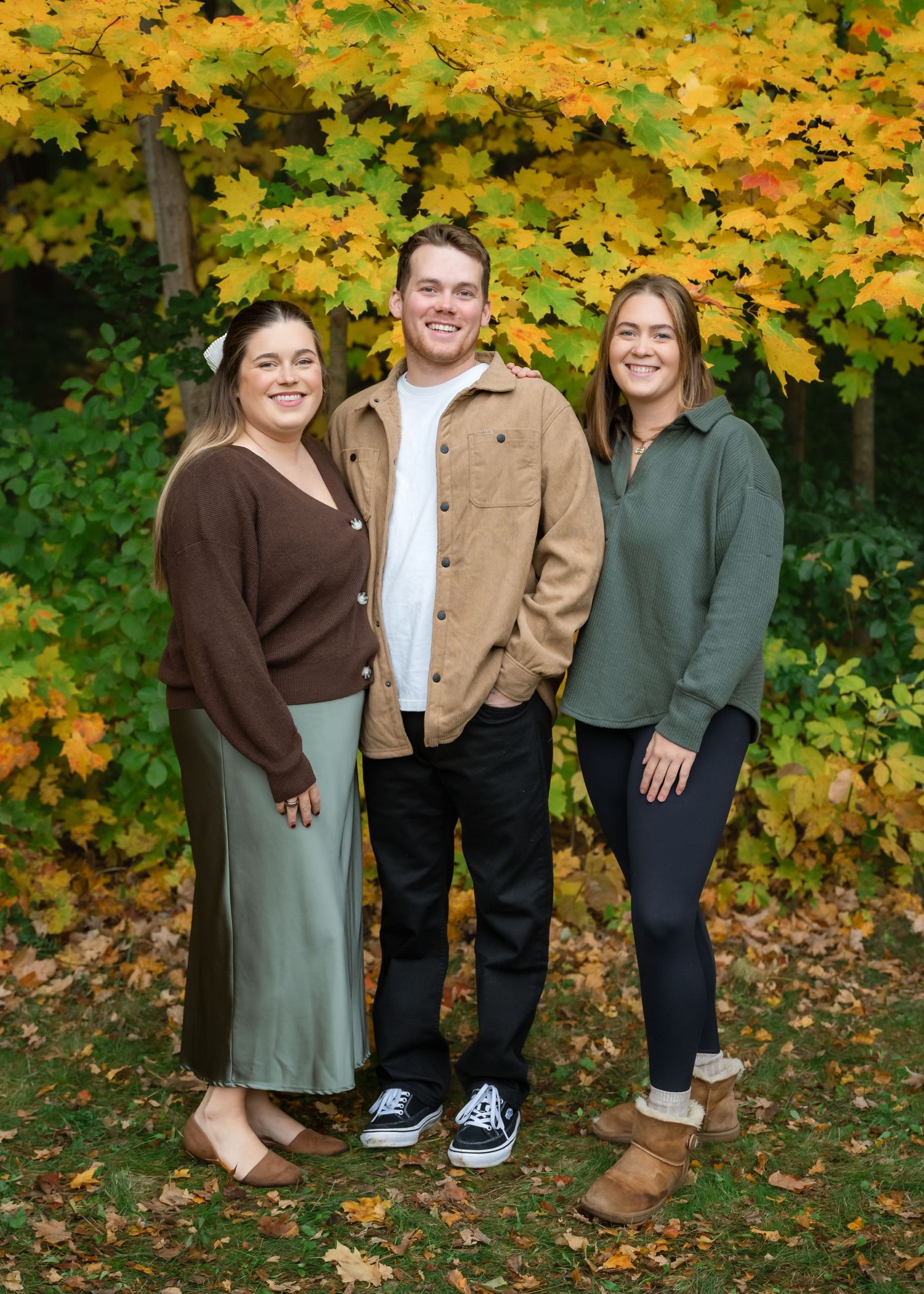 Three young adults smiling together outdoors among vibrant fall foliage with yellow and orange maple leaves.