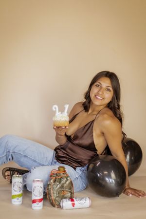 Young woman sitting on floor holding a cupcake with 21 candles, surrounded by balloons and White Claw cans in roy wa mini photography studio