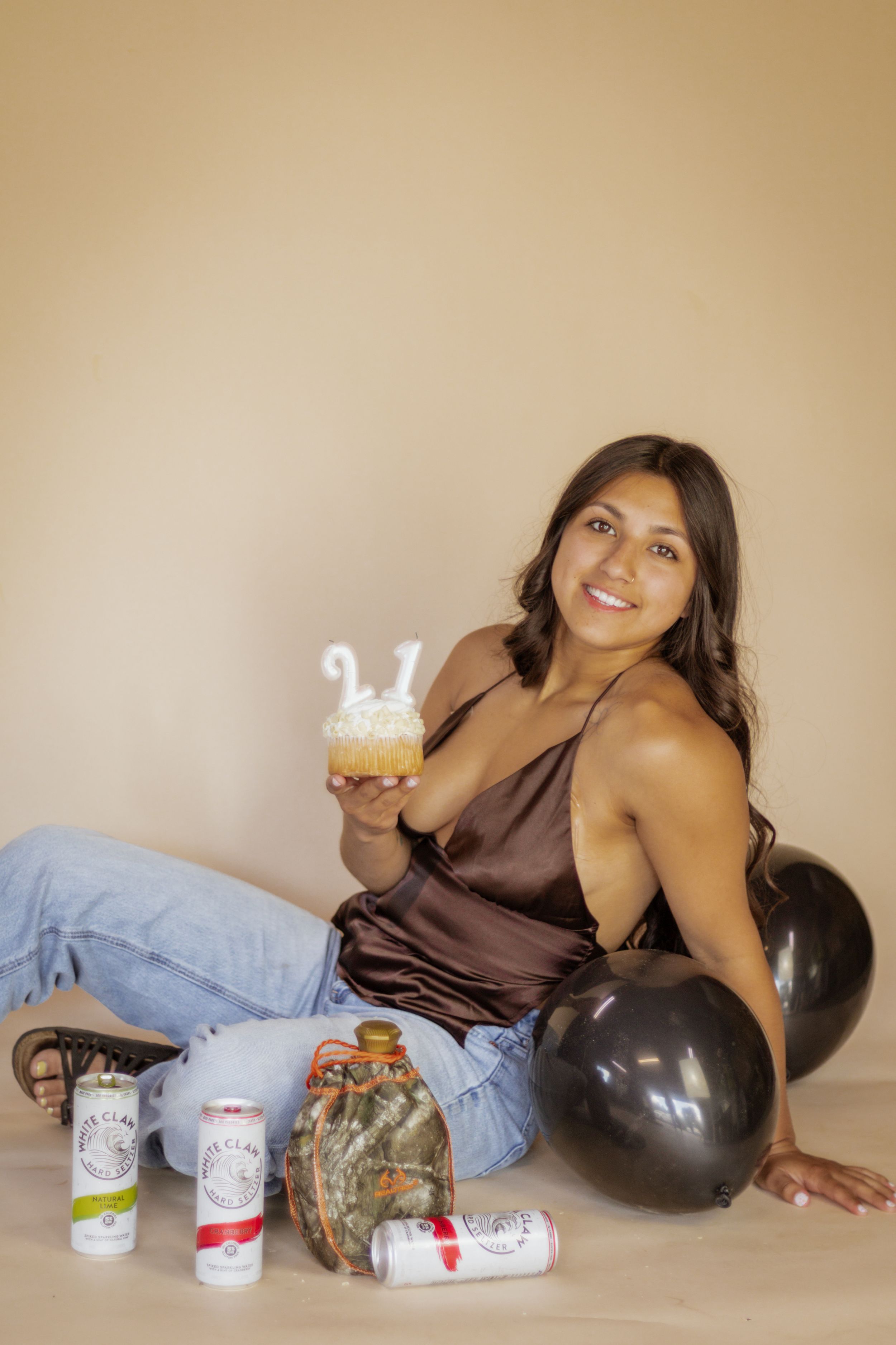 Young woman sitting on floor holding a cupcake with 21 candles, surrounded by balloons and White Claw cans in roy wa mini photography studio