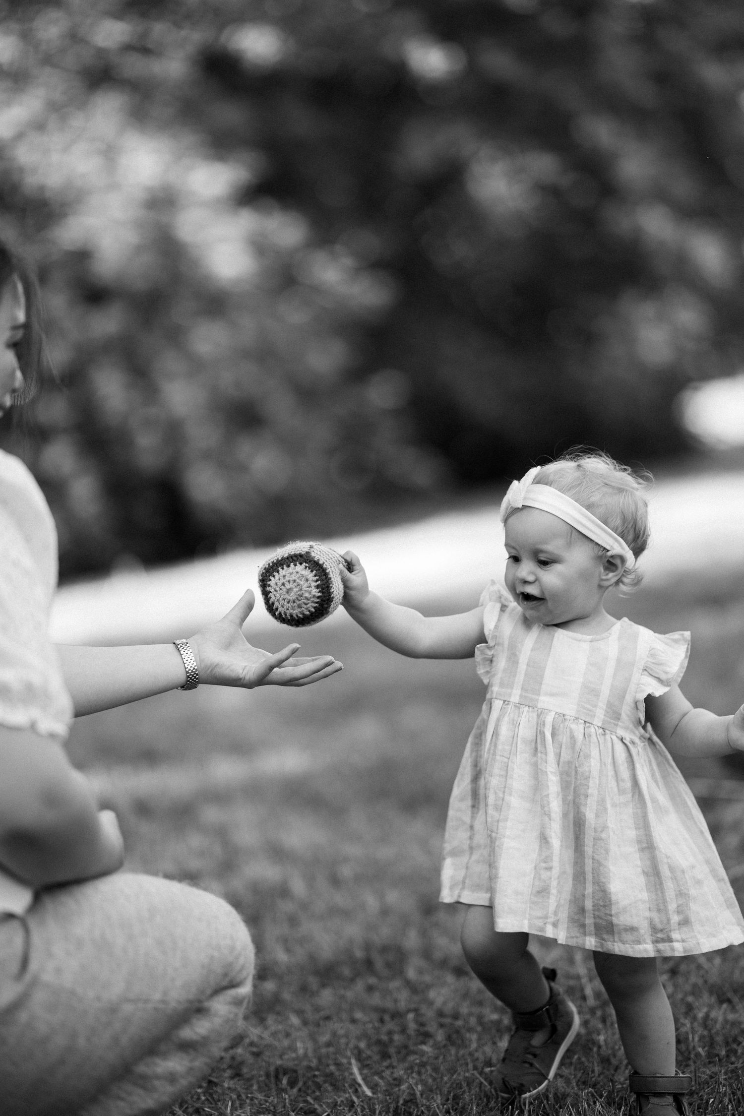 Ett litet barn kommer gåendes i gräs och räcker en virkad boll till sin mamma som sitter på knä och sträcker ut sin hand.