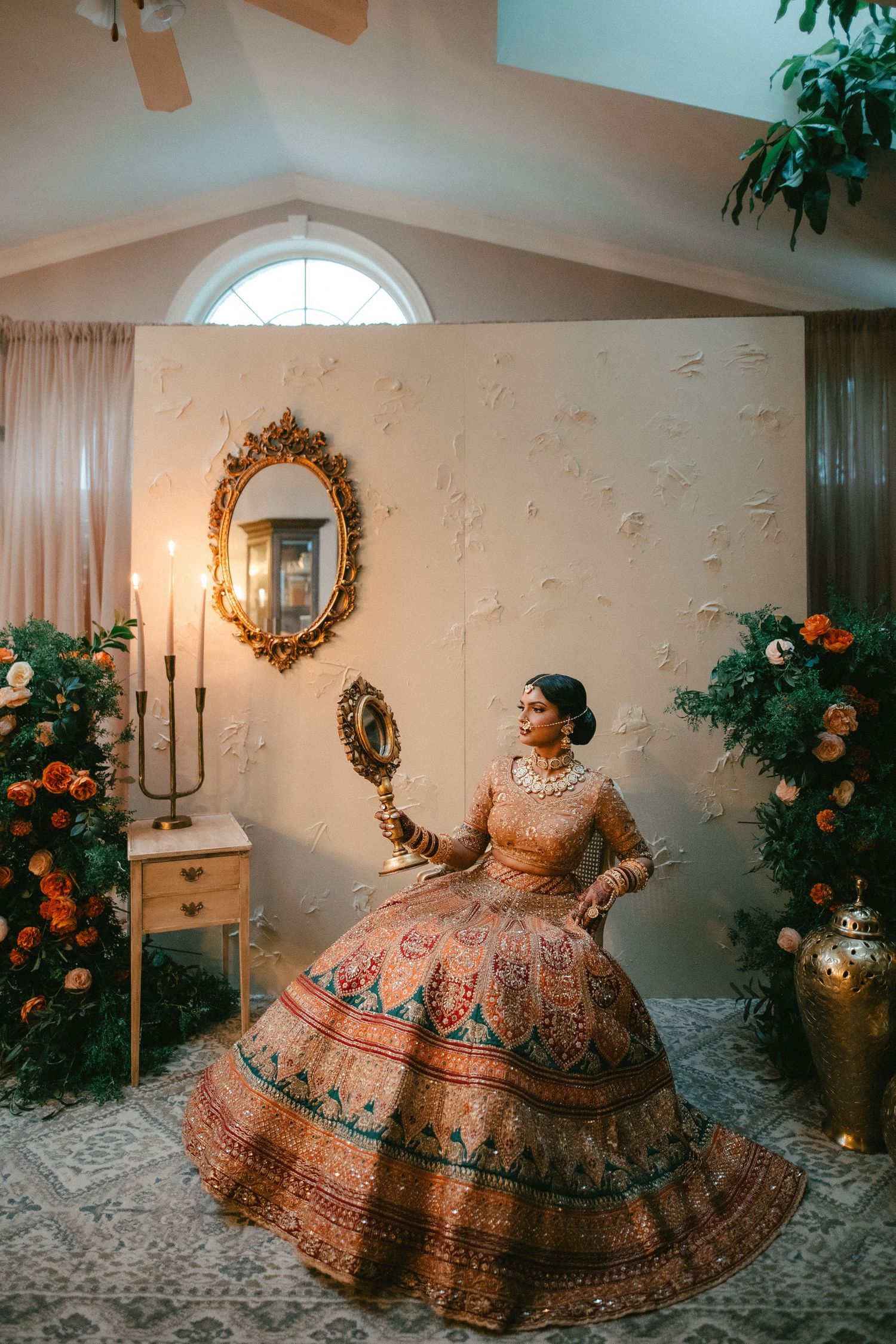 Indian bride in ornate lehenga holding mirror, surrounded by floral arrangements and golden decor in elegant bridal setting.