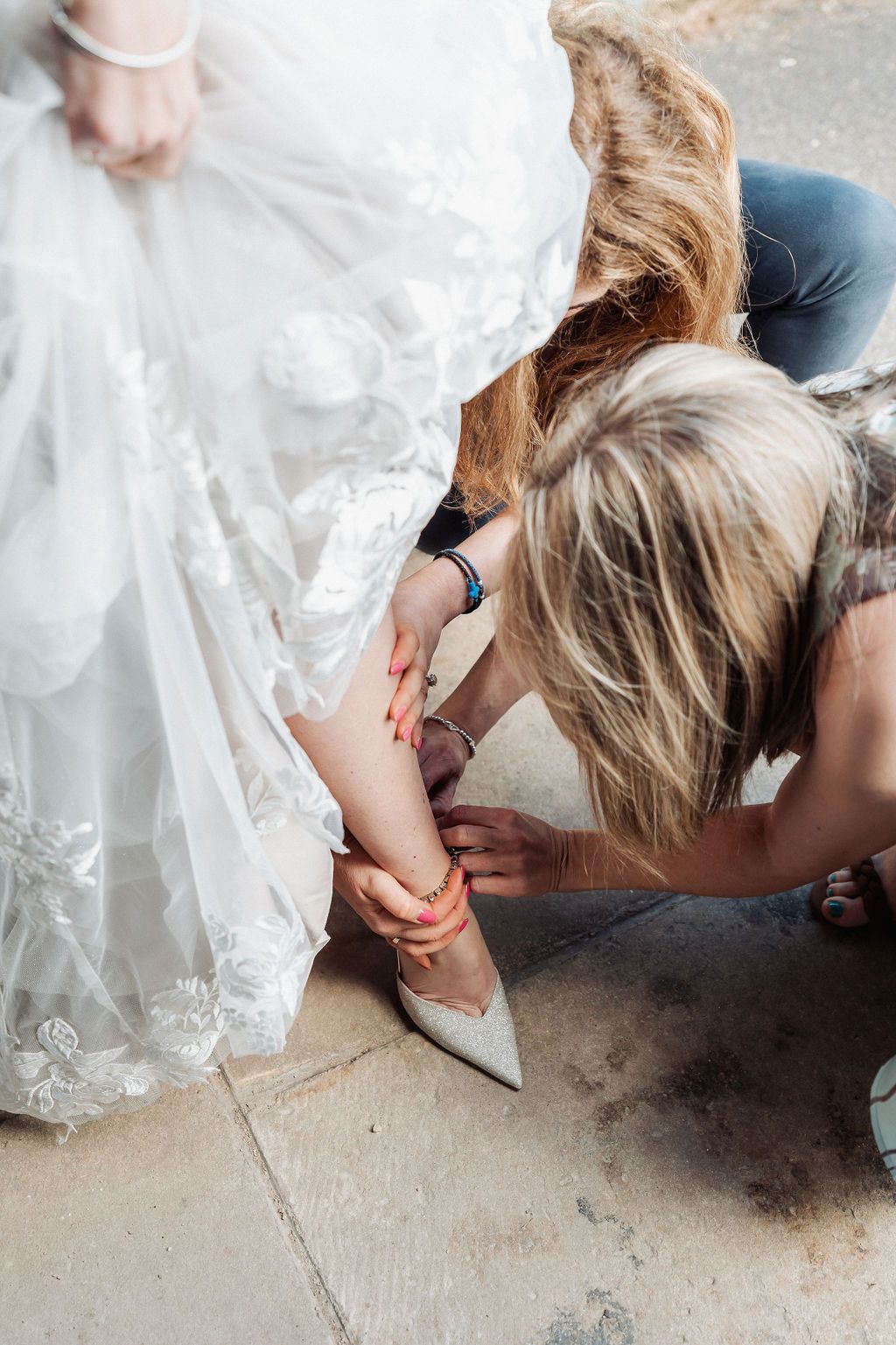 Bridesmaid helping bride put on white heeled wedding shoe, adjusting ankle strap on lace bridal gown.