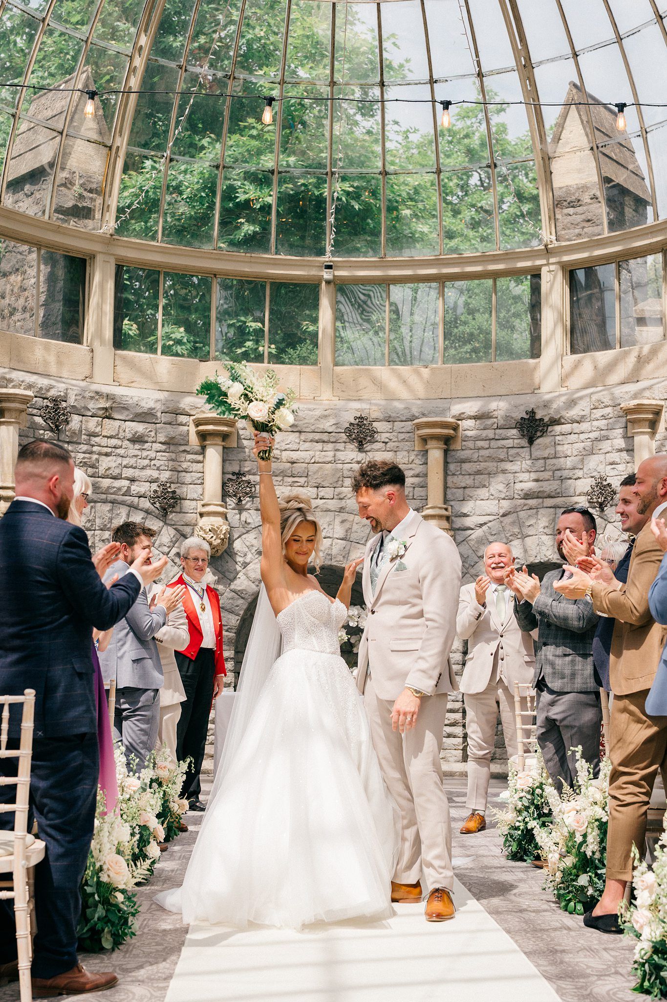 Bride and groom celebrate at wedding ceremony in elegant glass-domed stone venue, surrounded by cheering guests and floral arrangements.