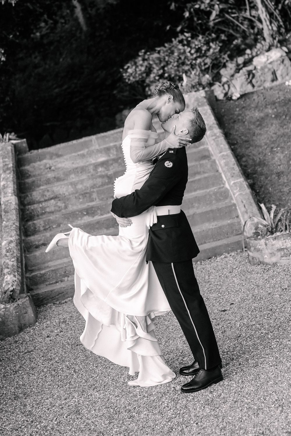 Black and white photo of a groom in military uniform lifting and kissing his bride in a wedding gown outdoors.