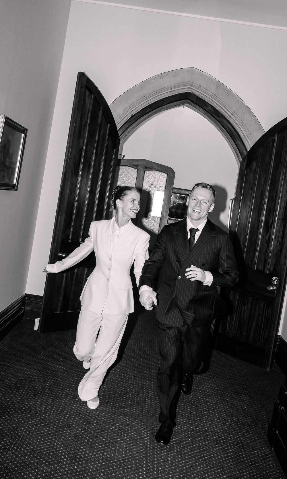 Black and white photo of a joyful couple in wedding attire running hand in hand through an arched doorway.