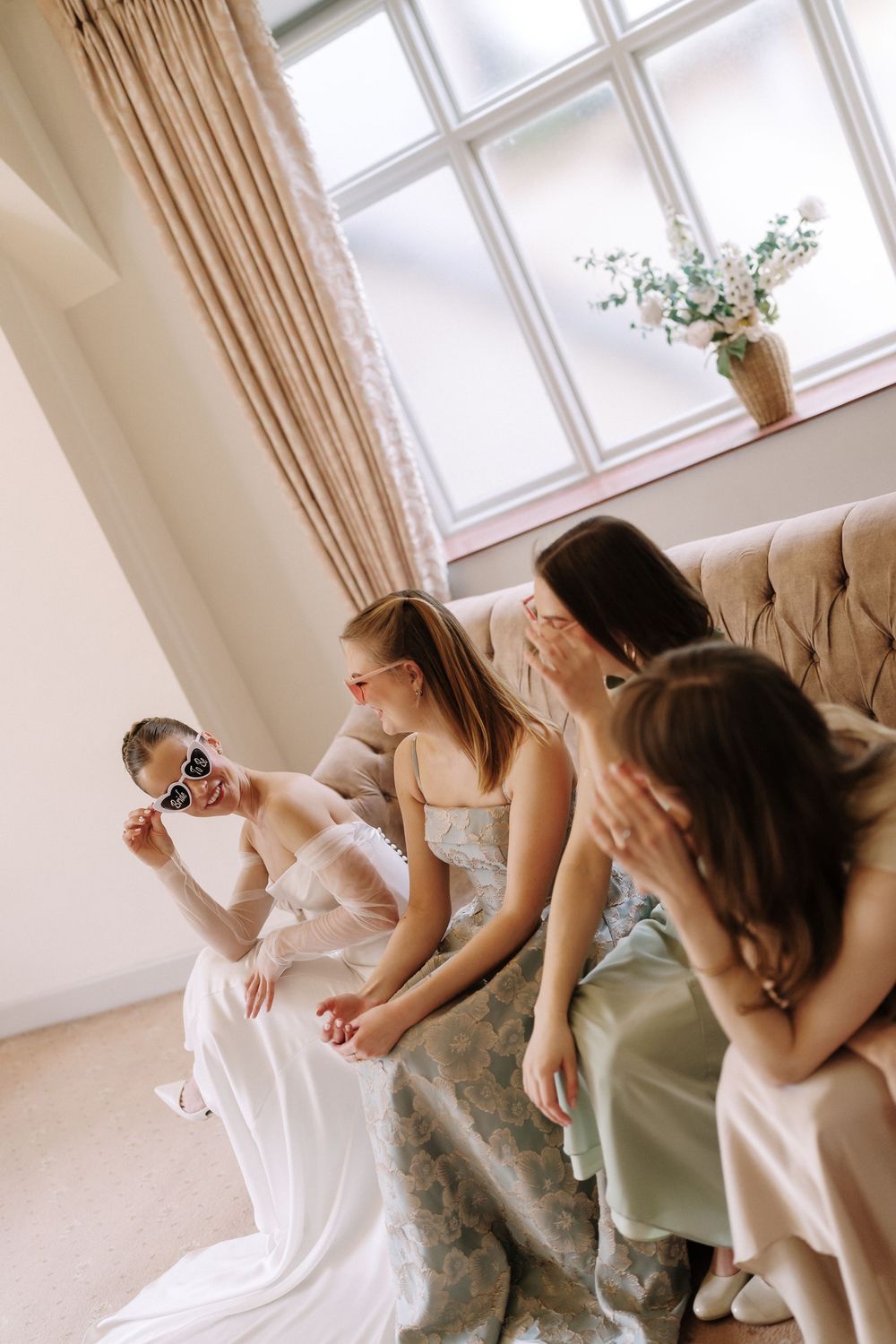 A bride wearing sunglasses laughs with three bridesmaids in satin dresses seated on an elegant sofa.