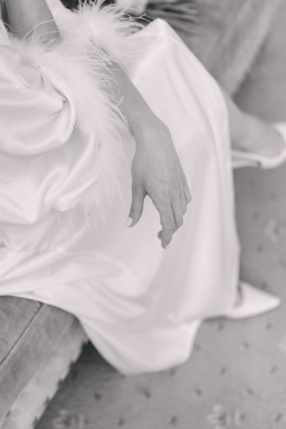 Black and white photo of a bride in a white satin gown with feather trim, her hand resting gracefully on her lap.