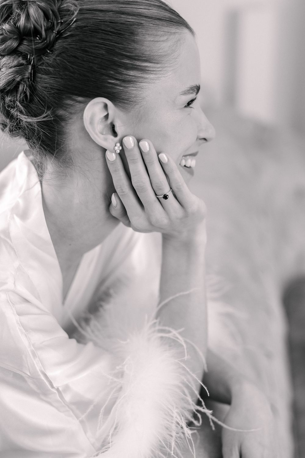 Smiling bride in white robe touches her face, showcasing floral earrings and engagement ring, in black and white photo.