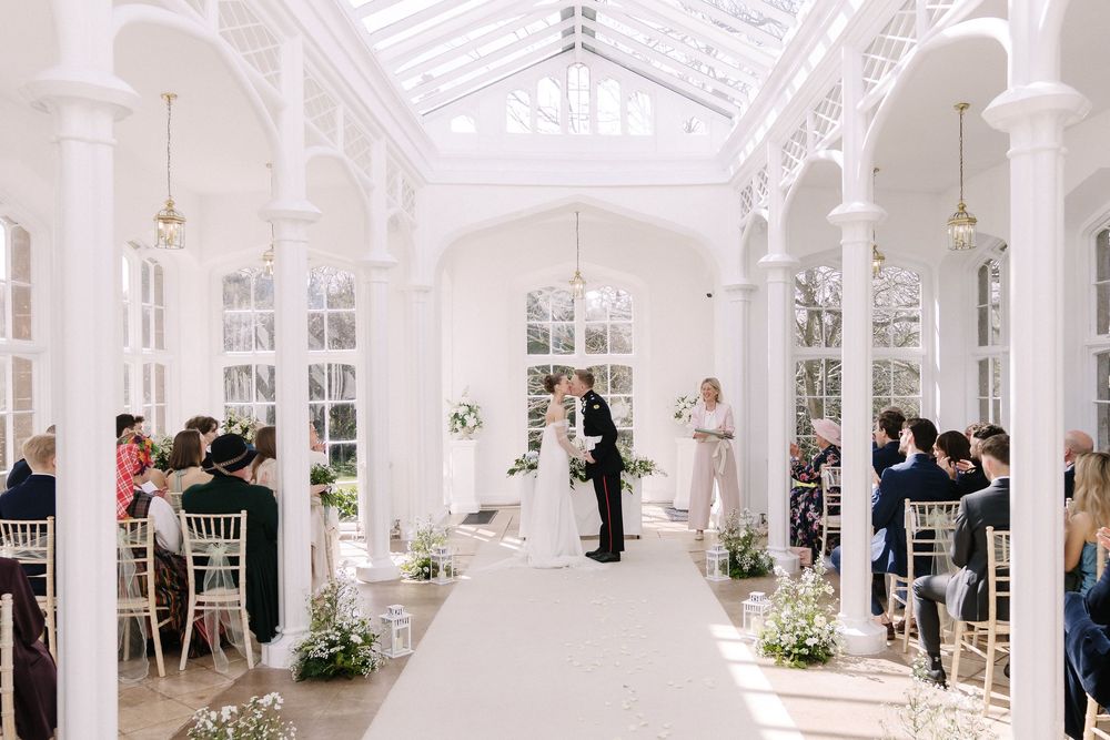 Elegant wedding ceremony in a bright white Victorian conservatory with floral arrangements and guests seated in chiavari chairs.