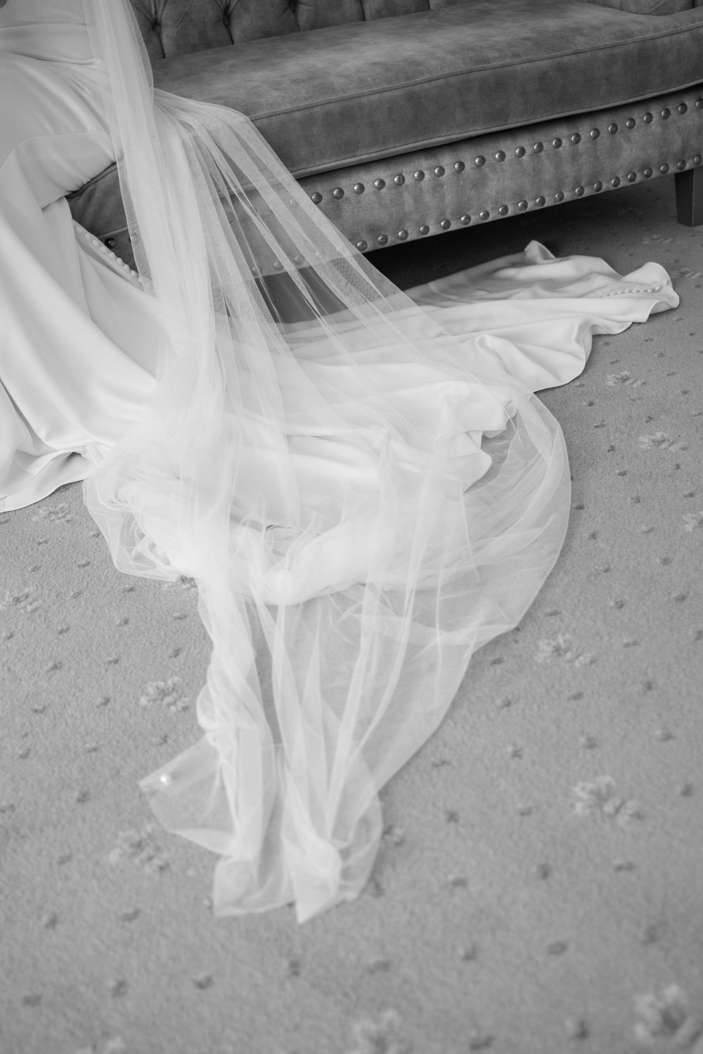 Black and white photo of a flowing white bridal veil and wedding dress train spread across a carpeted floor.