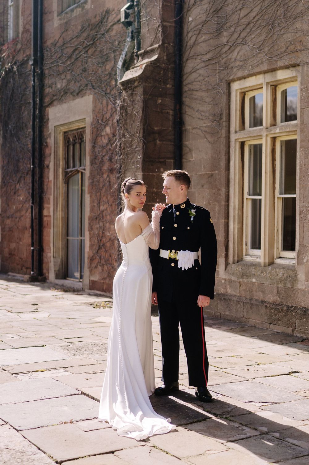 Bride in white gown and groom in military dress uniform pose together outside a historic stone building.