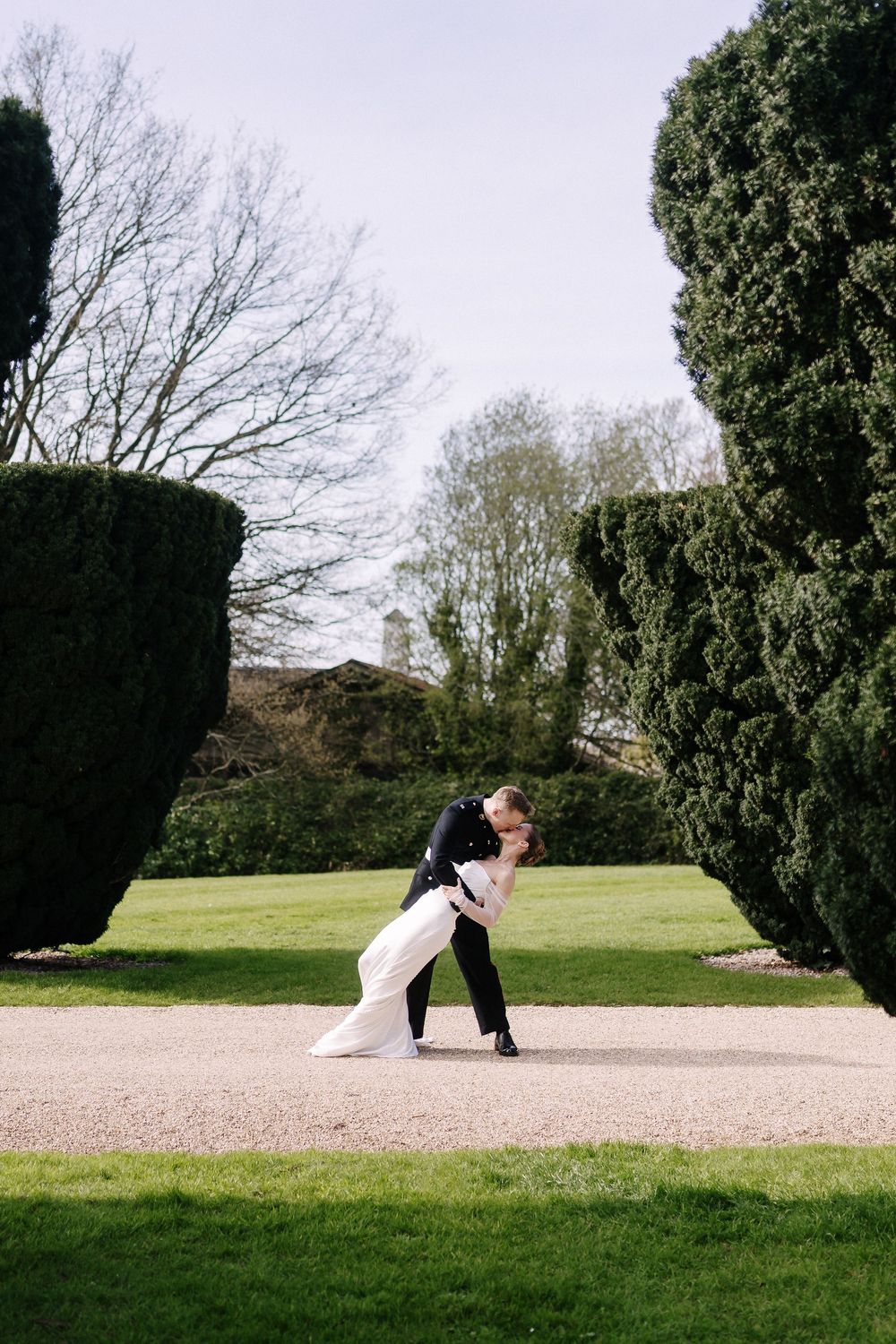 Bride and groom share a romantic dip kiss on a gravel path surrounded by lush green topiary trees in an elegant garden.