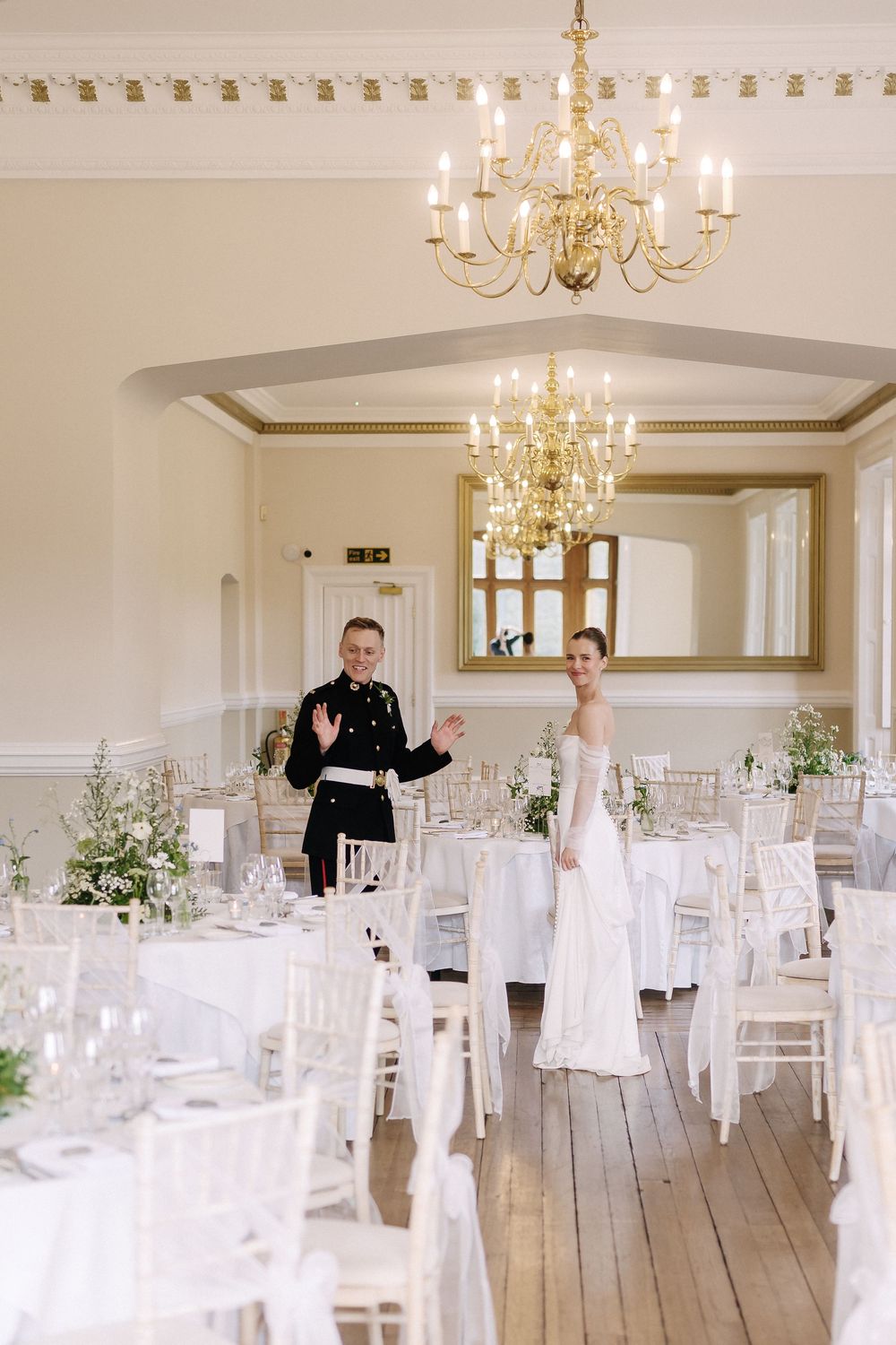Bride and groom in military uniform enter elegant white wedding reception hall with chandelier and floral centerpieces.