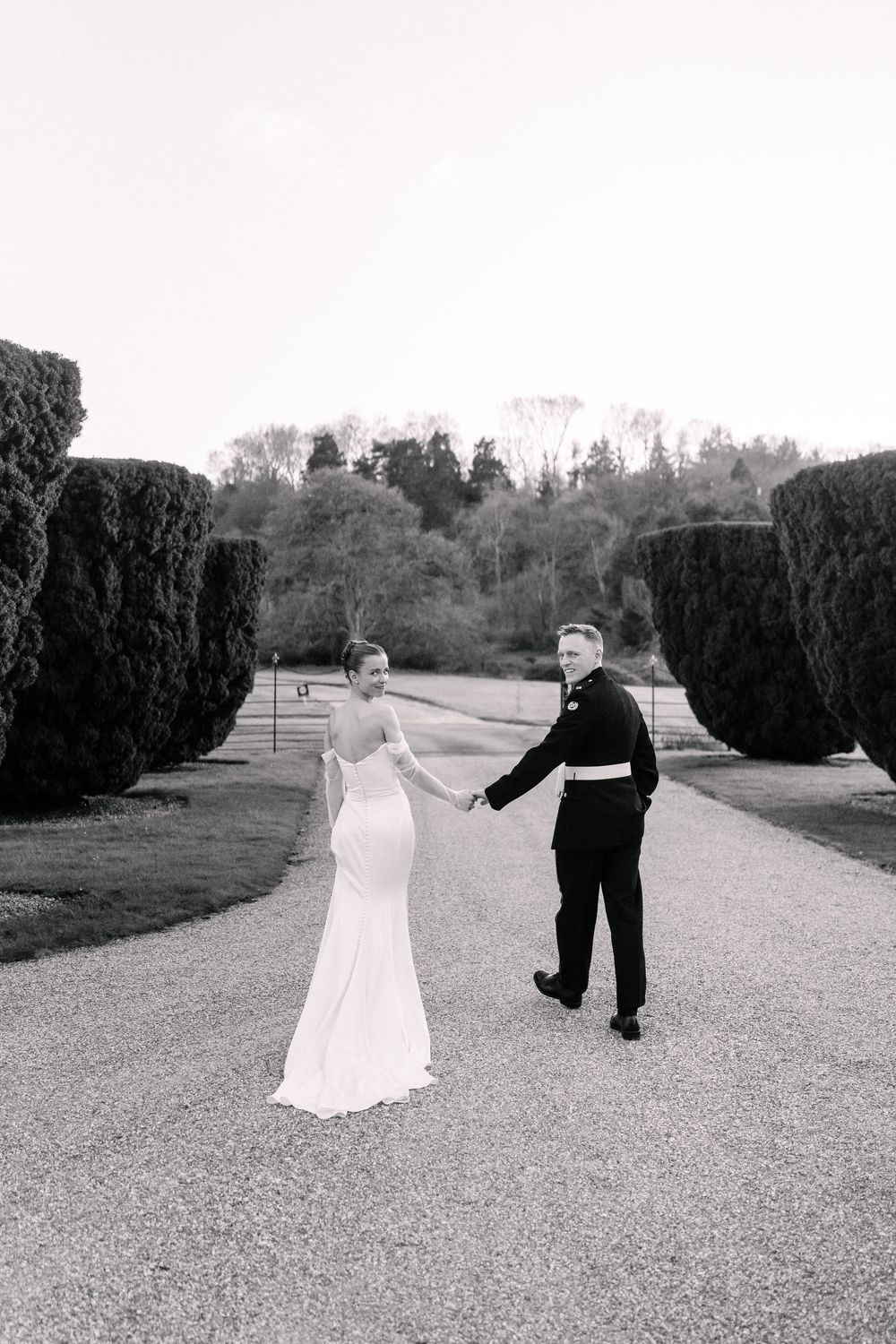 Bride and groom hold hands walking down grand garden path flanked by tall hedges in black and white photo.