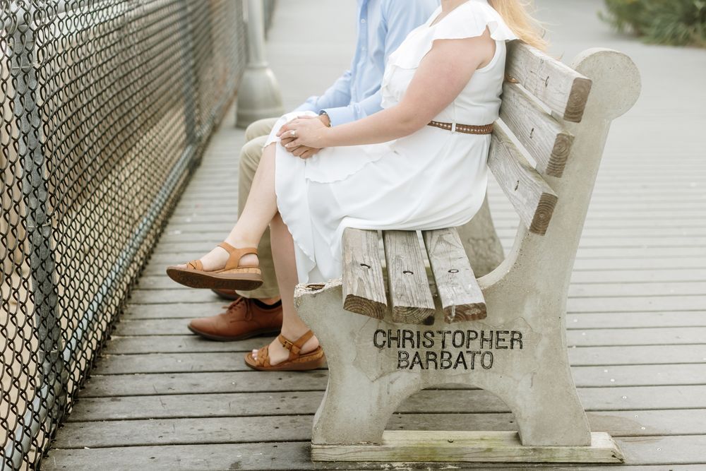 Couple sits on a memorial bench dedicated to a man on a wooden boardwalk in Point Pleasant New Jersey. Seacoast NH engagement photographer.