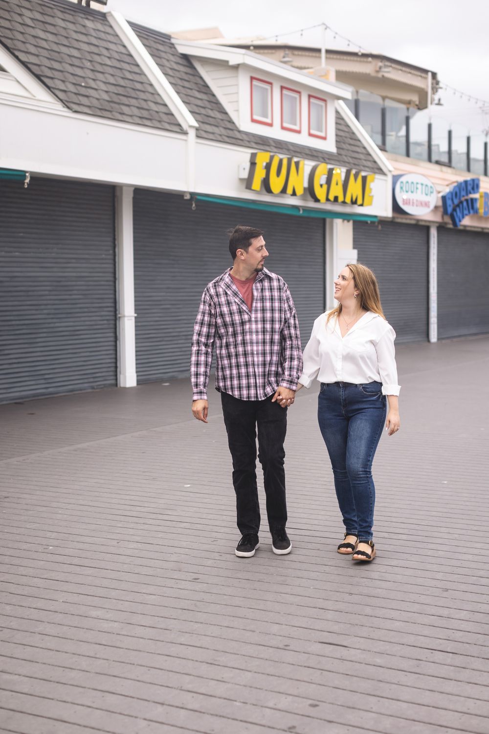 Couple walks down boardwalk holding hands with metal gates closed on beach boardwalk games. Old Orchard Beach engagement photographer.
