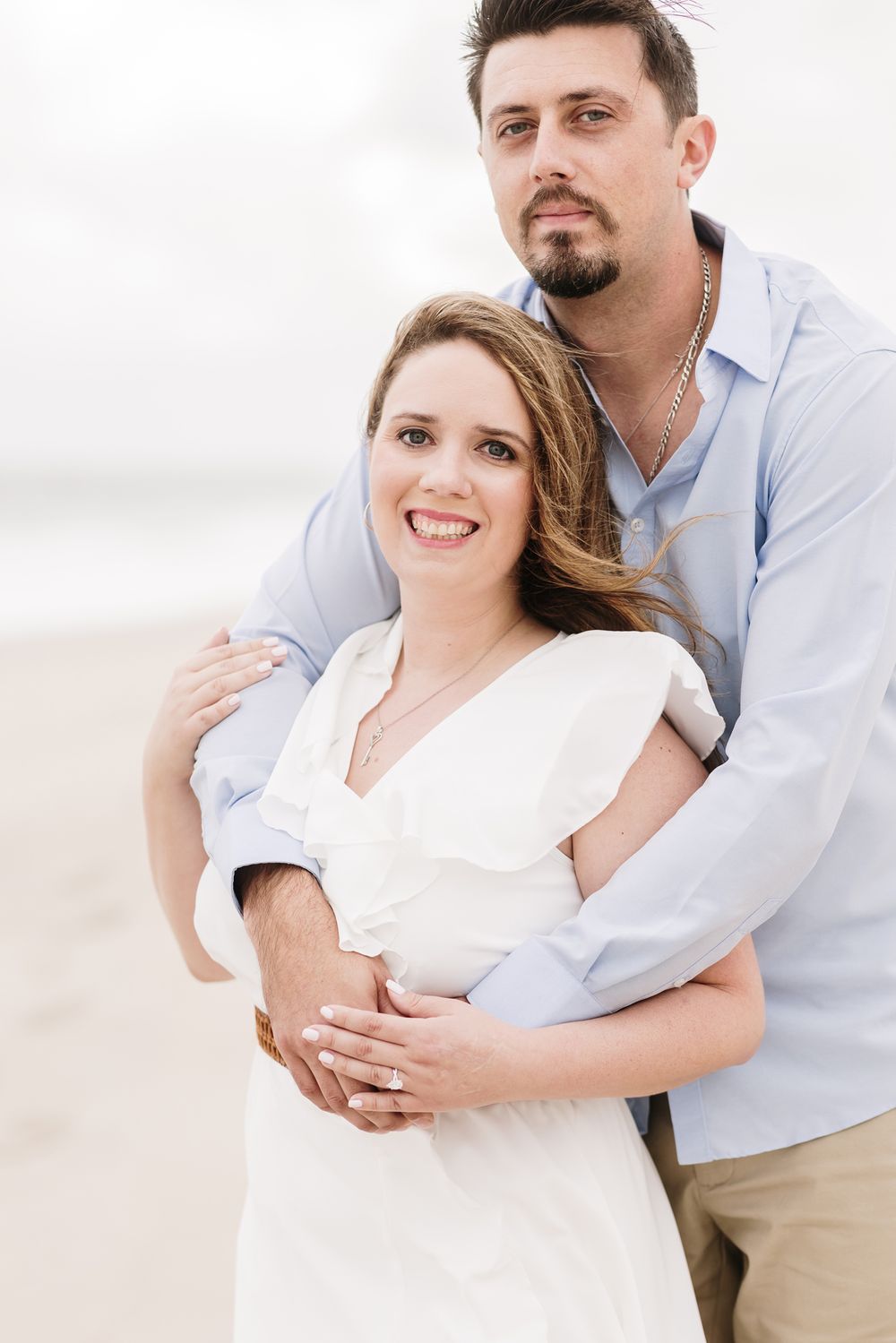 Woman in white dress is held by fiancee in blue shirt for engagement photos on cloudy beach day. North Shore engagement photographer.