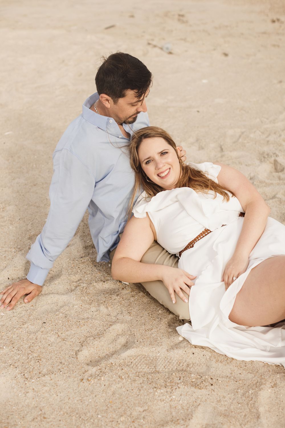 Woman in white dress lays on man's lap on beach for engagement photos as he looks down at her. Portland Freeport engagement photographer.