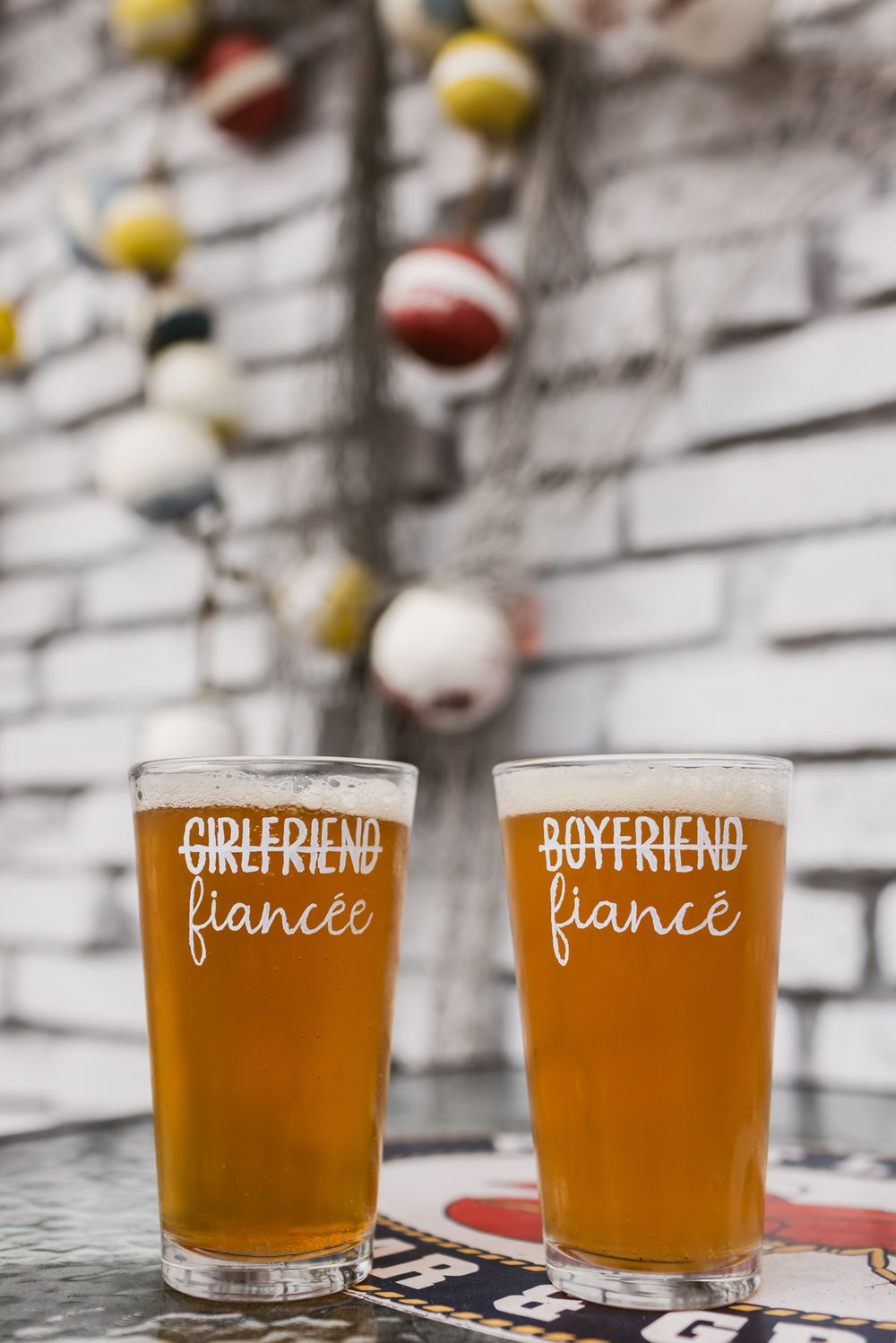 Two pint glasses engraved with strikethrough Girlfriend/Boyfriend and fiancee/fiance, filled with beer on a table outside restaurant.