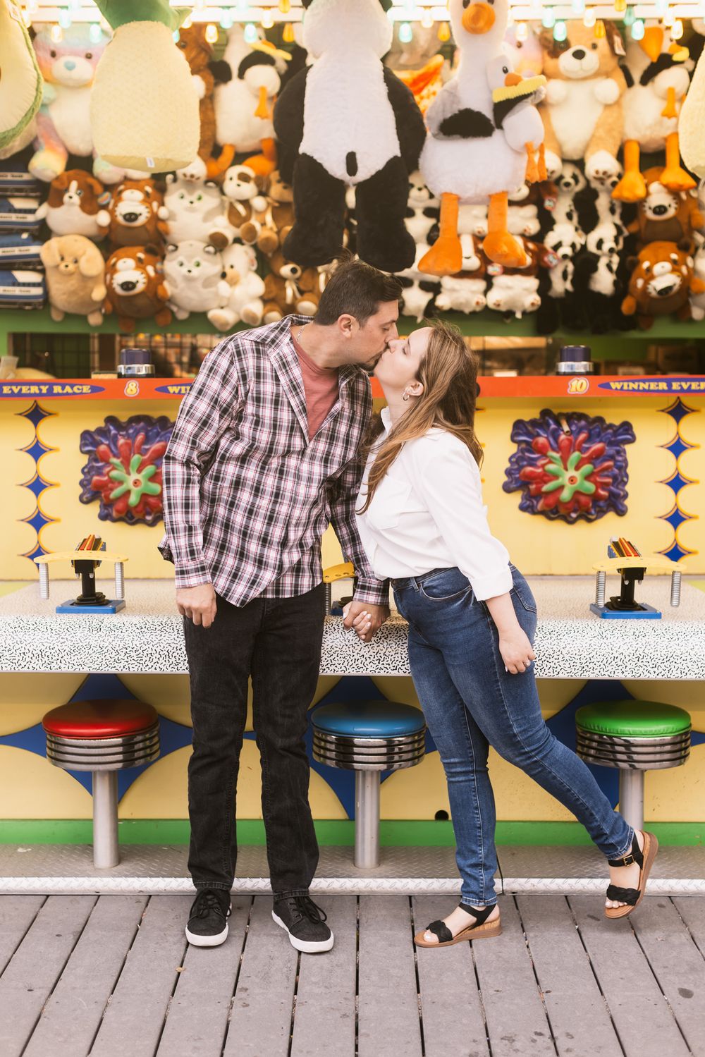 Woman in white shirt and jeans leans to kiss fiancee in plaid shirt with stuffed animals in background at boardwalk game.