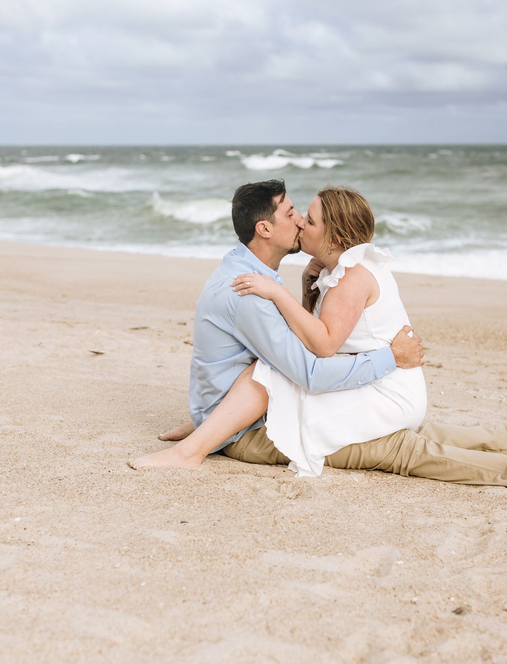 Couple sits with legs crossed into each other for engagement photos on sunny beach day. Rockport Gloucester engagement photographer.