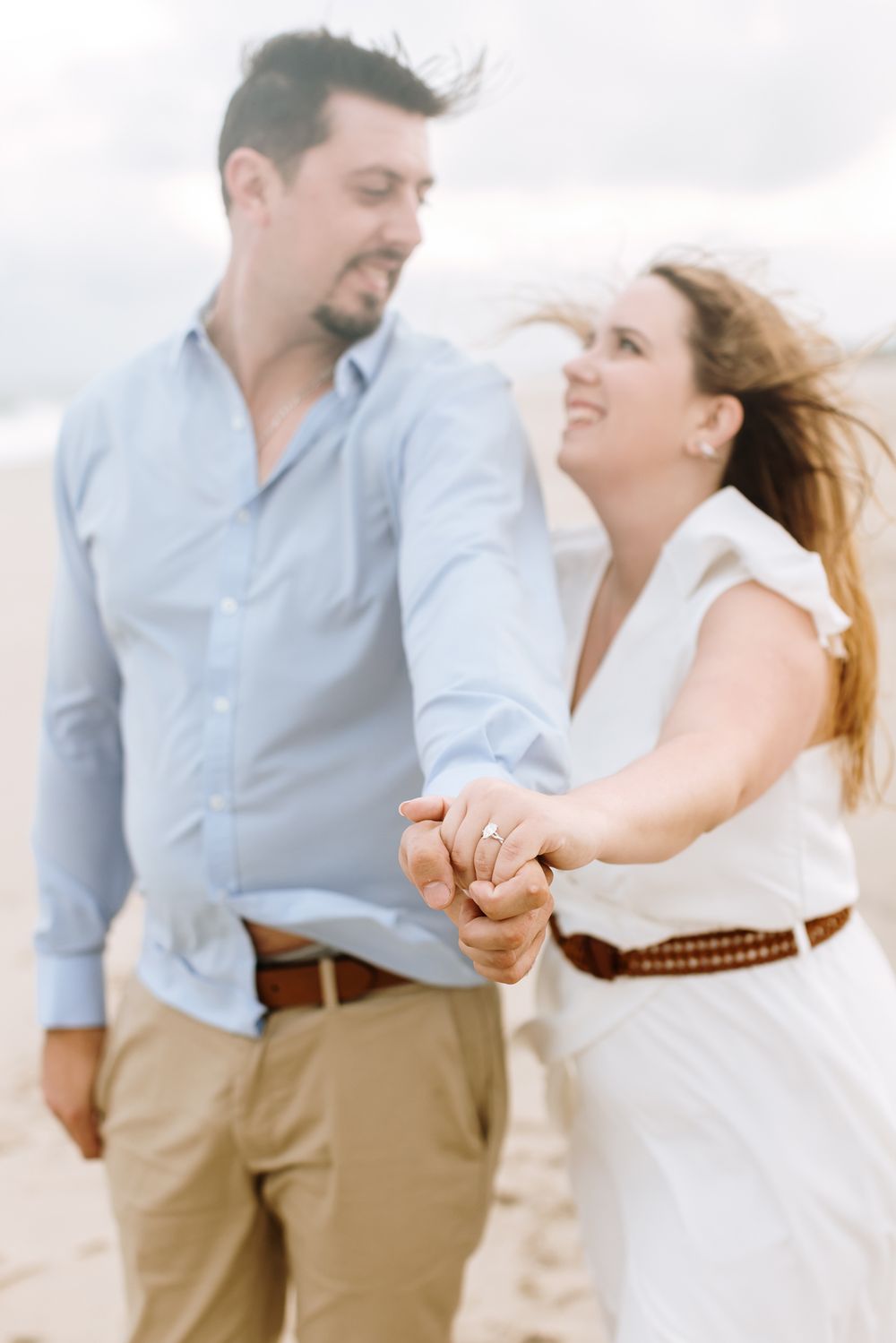 Focus on engagement ring of couple's hands clasped with couple smiling in background for engagement photos. Harvard engagement photographer
