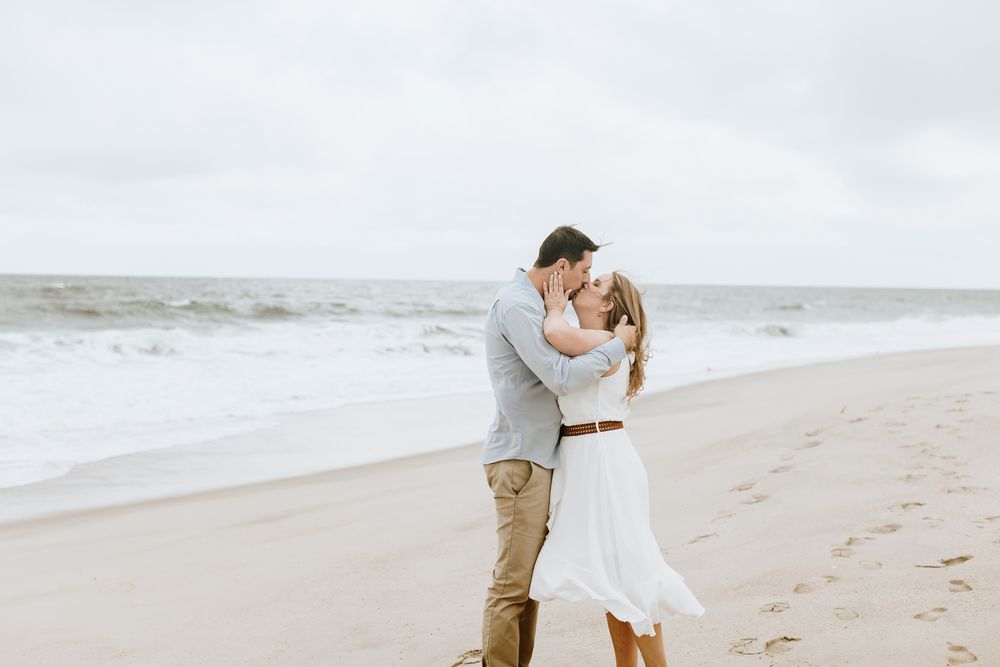 Man in blue shirt and tan khakis kisses woman in white dress flowing in wind on beach for engagement photos. Concord engagement photographer