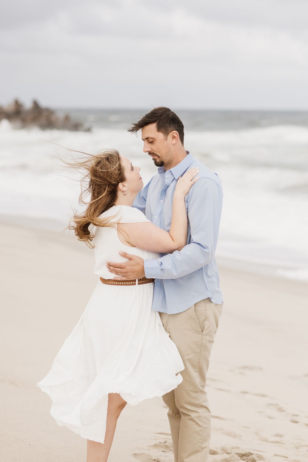 Woman in white dress is embraced by man in blue shirt and tan khakis on windy, cloudy beach day. Monadnock NH engagement photographer