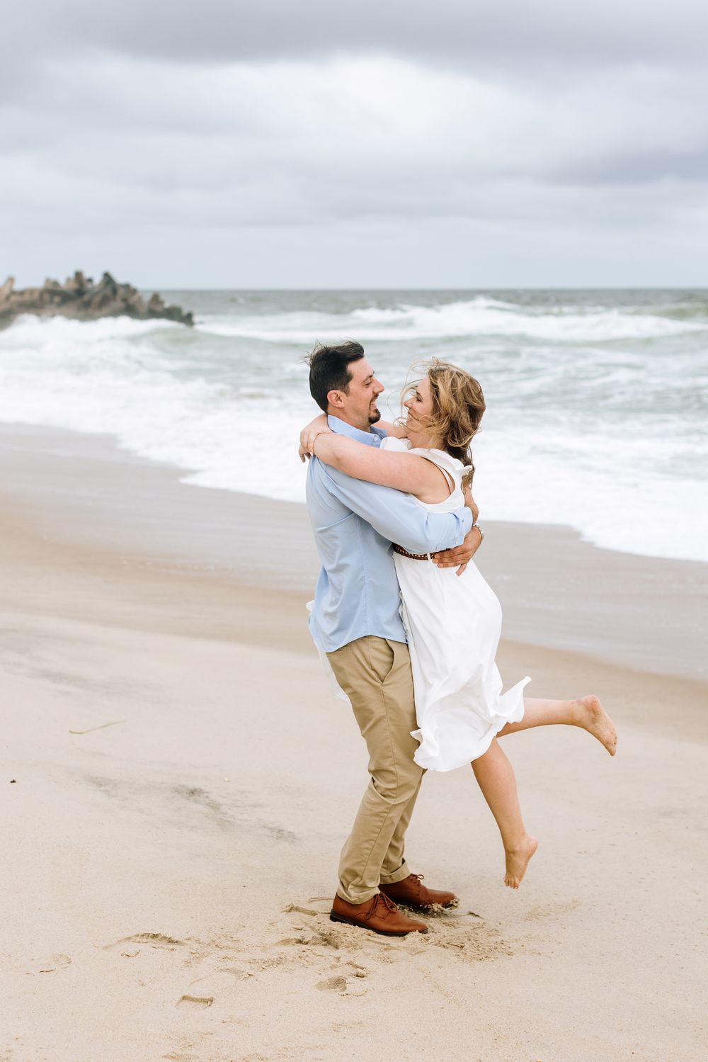 Man in blue shirt spins lady in white dress around on cloudy day beach for engagement photos. Southern Maine York engagement photographer.