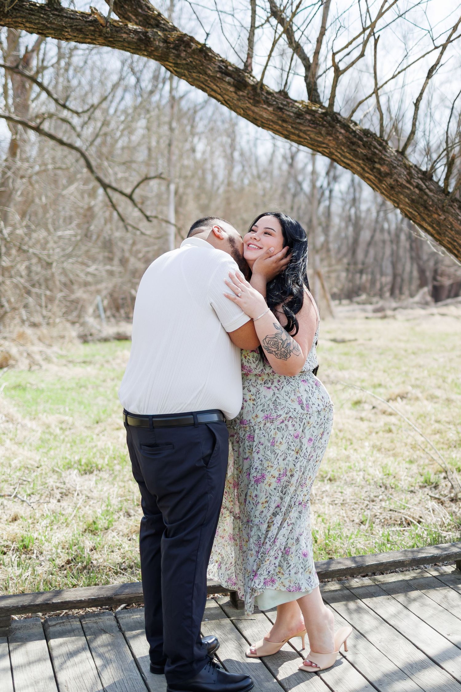 A young couple smiling and posing outdoors near a stone wall surrounded by lush green trees.