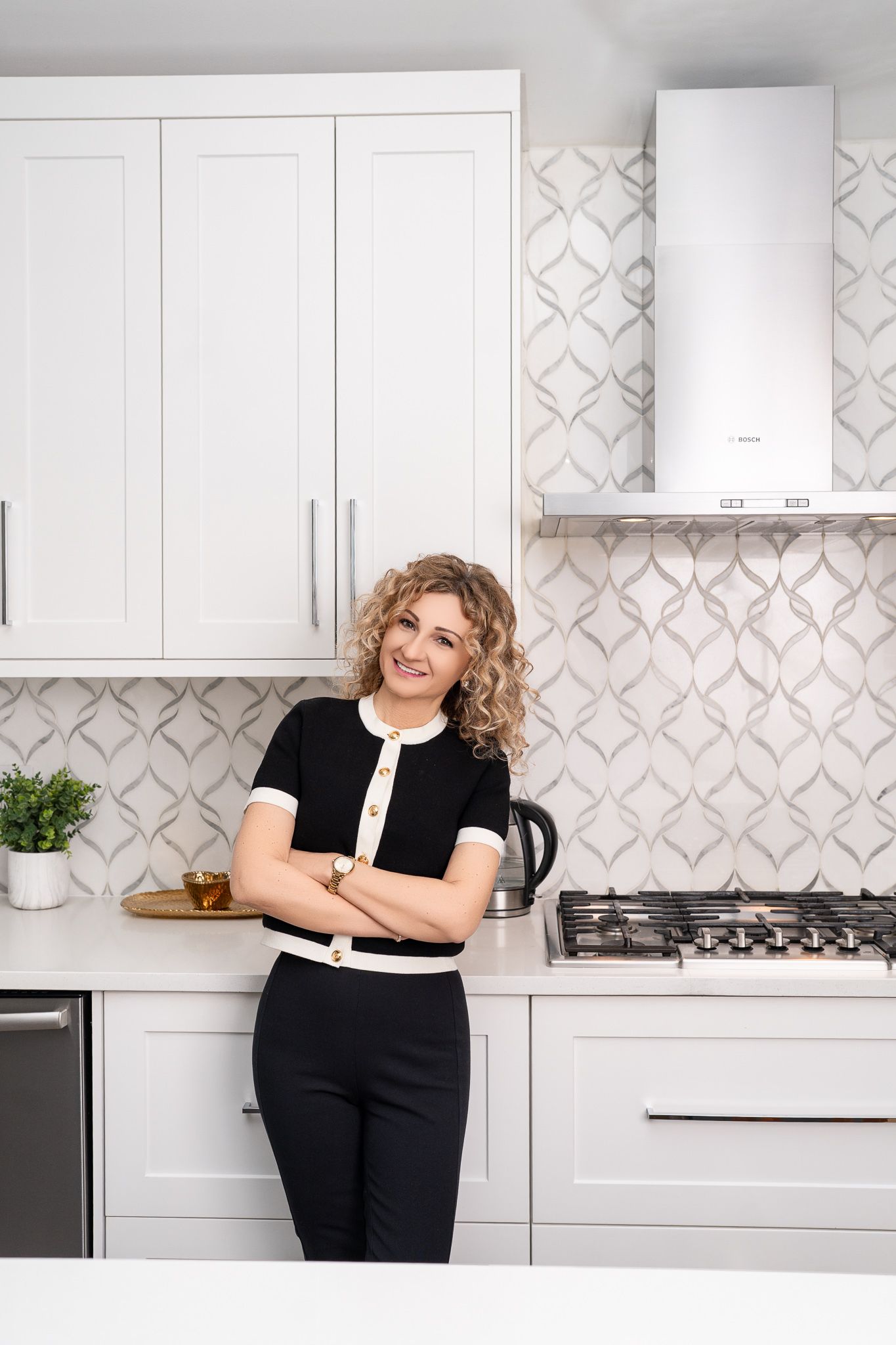 Smiling woman with curly hair in black outfit leaning against white kitchen counter with patterned tile backsplash.