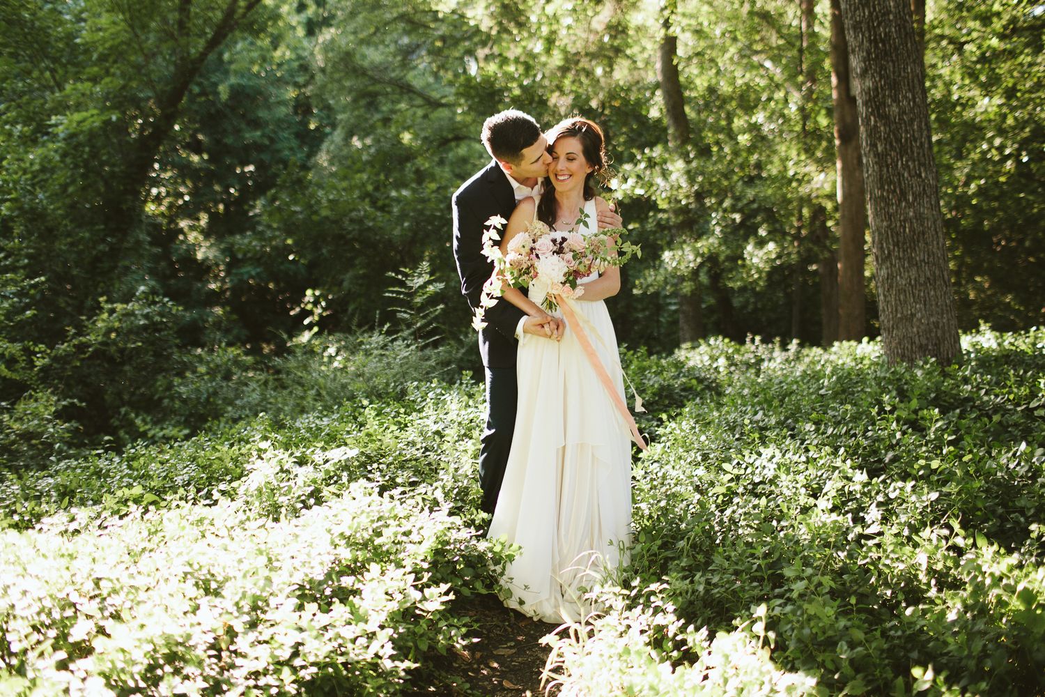 A bride and groom share a romantic moment in a sunlit forest, surrounded by lush greenery and wildflowers.