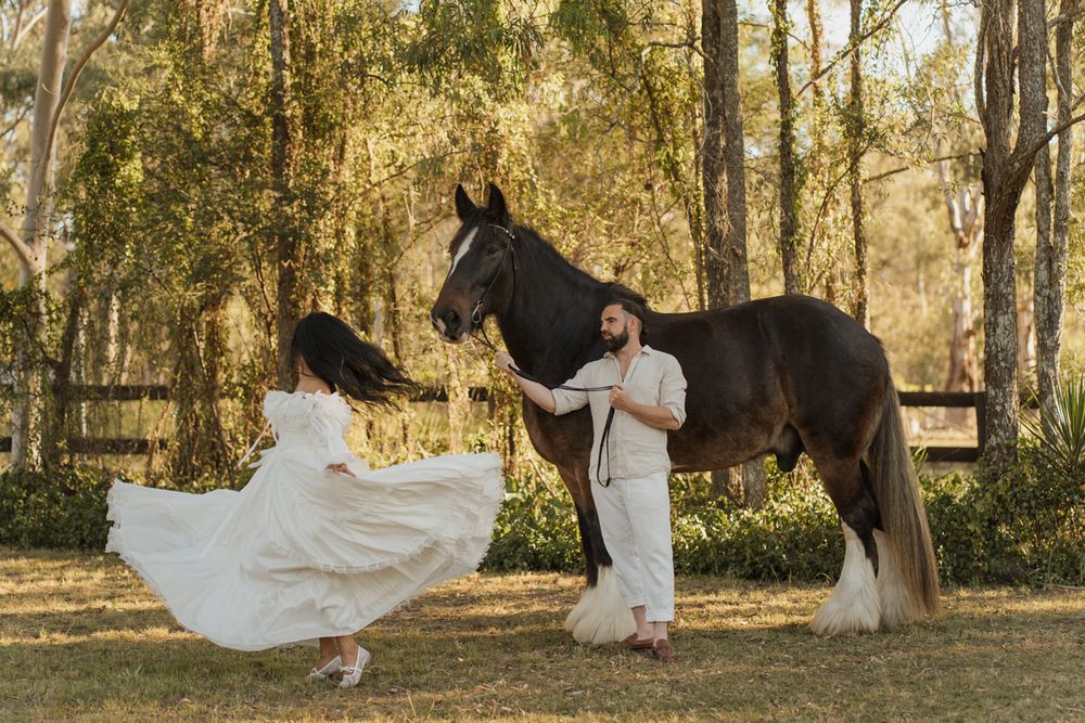 Couple in white attire dancing next to a dark horse in sunlit outdoor setting surrounded by trees during a photoshoot with a horse in sydney