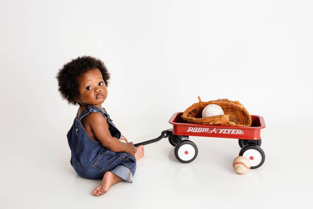 Toddler in denim overalls pulling a red Radio Flyer wagon filled with baseball glove and balls on white background.