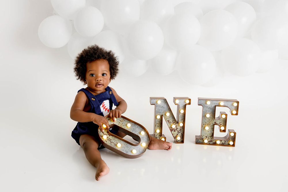 A baby sits smiling beside illuminated ONE marquee letters, celebrating their first birthday by Moreno Valley baby photographer.