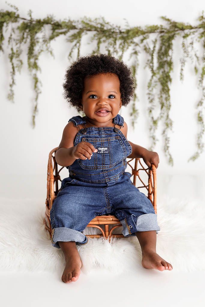 Smiling baby in denim OshKosh overalls sitting on a wicker chair by Moreno Valley baby photographer.