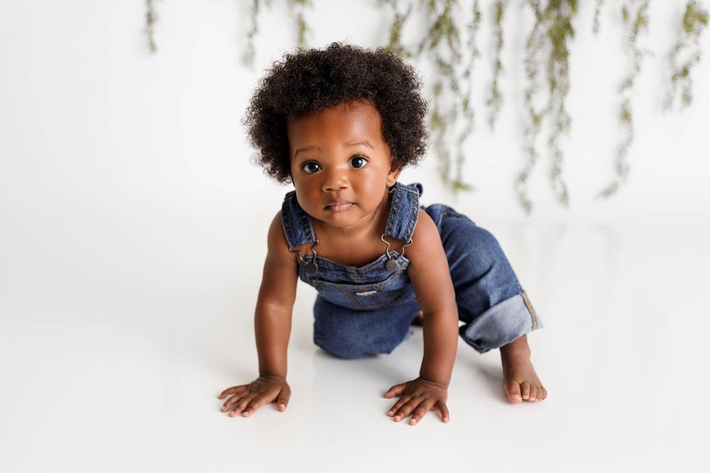 An adorable baby in denim overalls crawls on a white studio floor by Moreno Valley baby photographer.