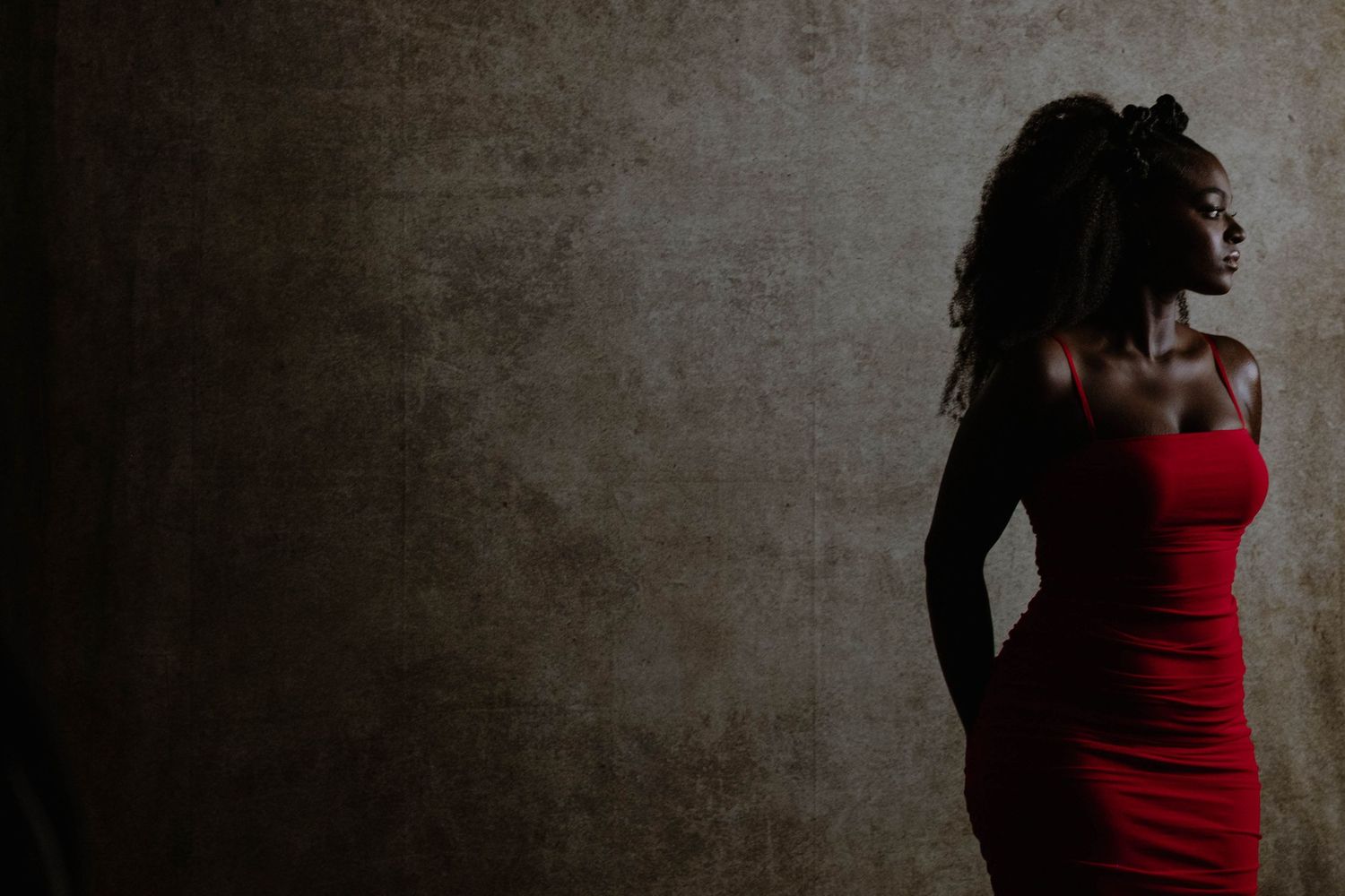 A Black woman in a red off-shoulder dress poses gracefully against a dark textured wall in dramatic lighting.