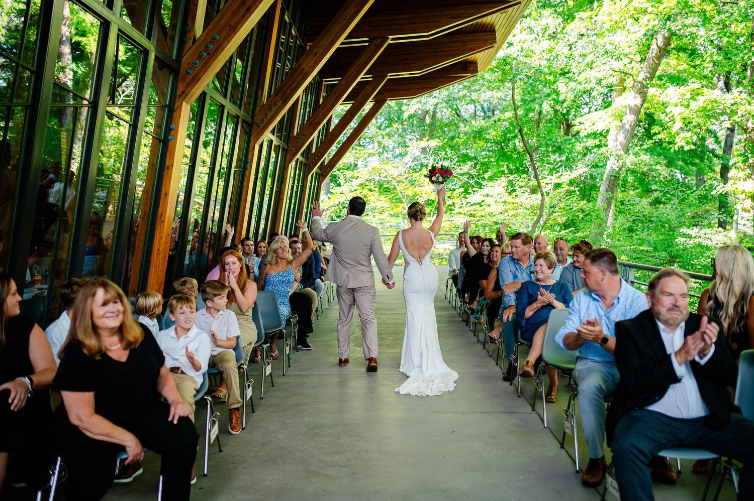 Bride and groom walk down the aisle at an outdoor woodland wedding venue as guests celebrate.