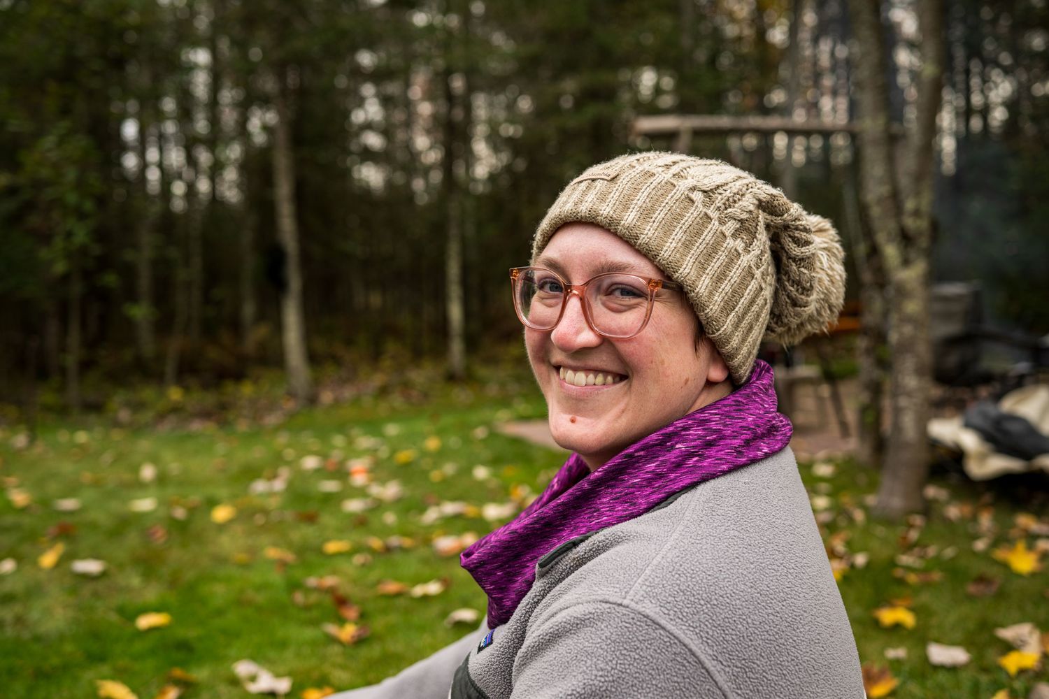 Smiling woman wearing a knit beanie and purple scarf outdoors in autumn with trees and fallen leaves.