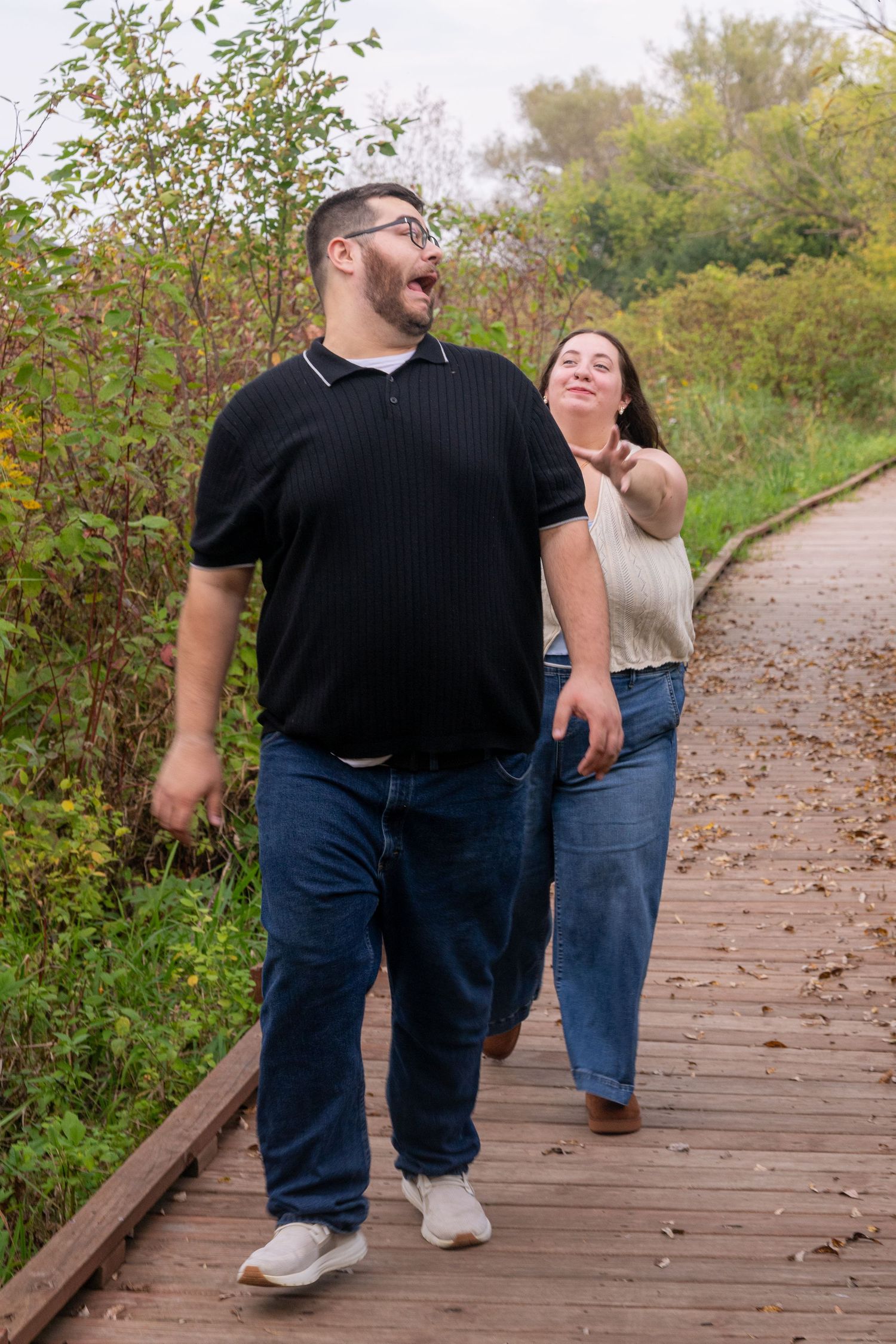 Couple laughing and walking together on a wooden boardwalk path surrounded by autumn foliage.